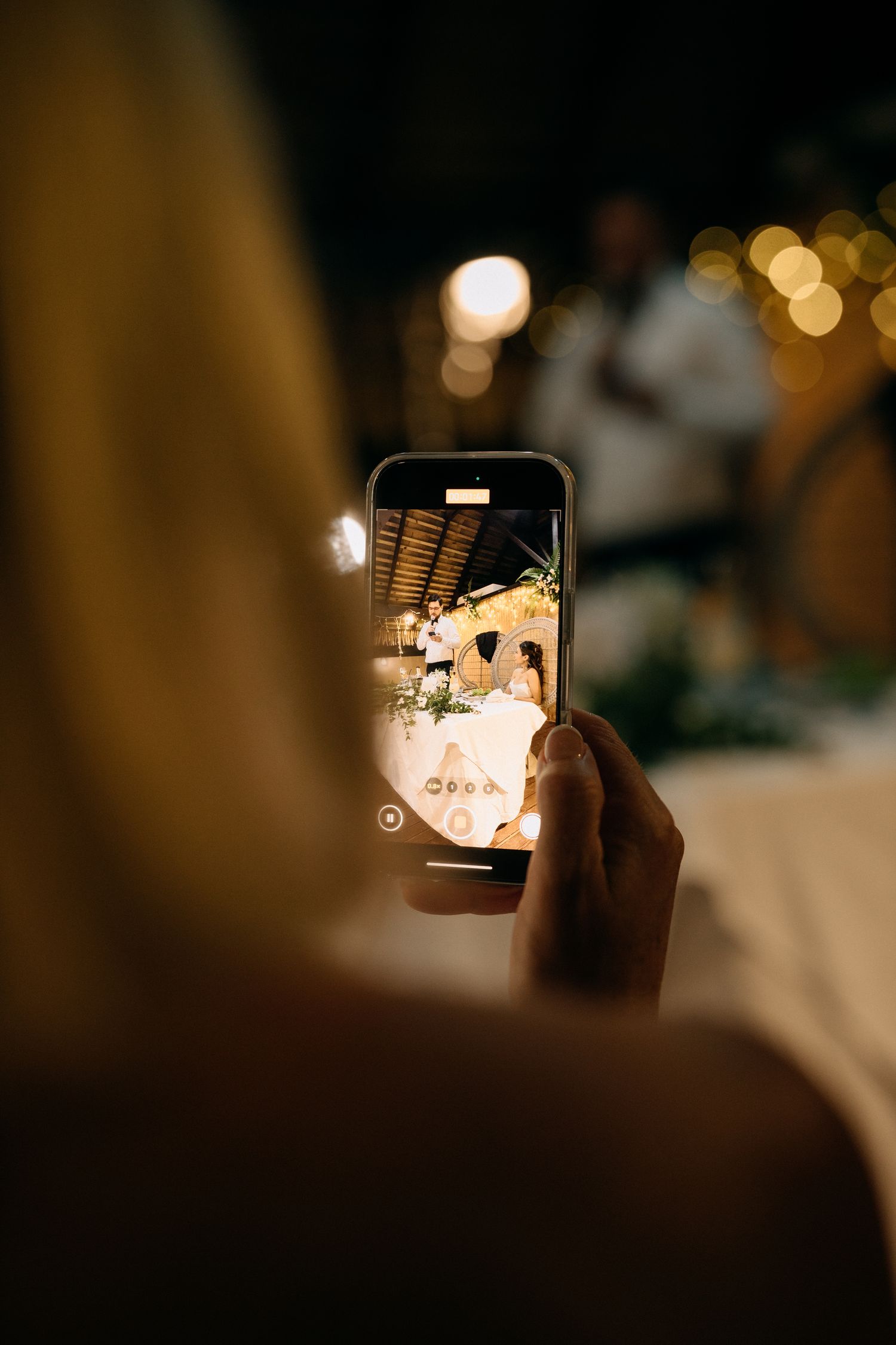 Guest capturing wedding couple on smartphone screen at elegant evening reception with warm bokeh lights.