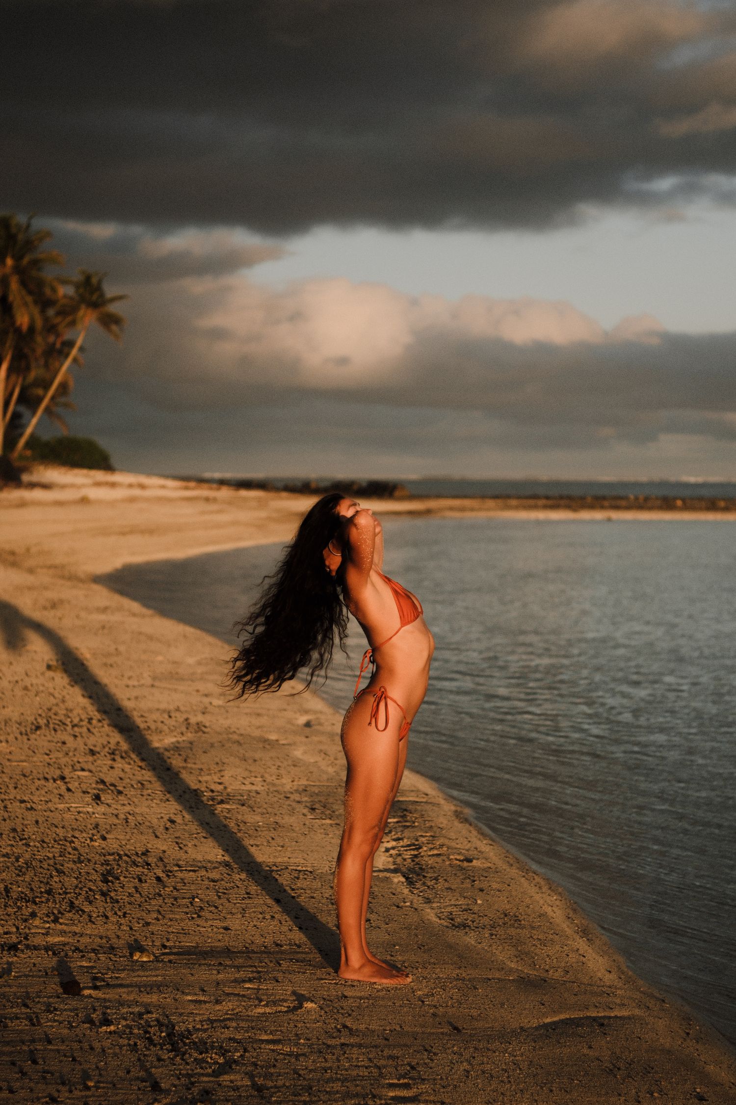 Woman in bikini posing on sandy beach at golden sunset with dramatic storm clouds and palm trees in background.