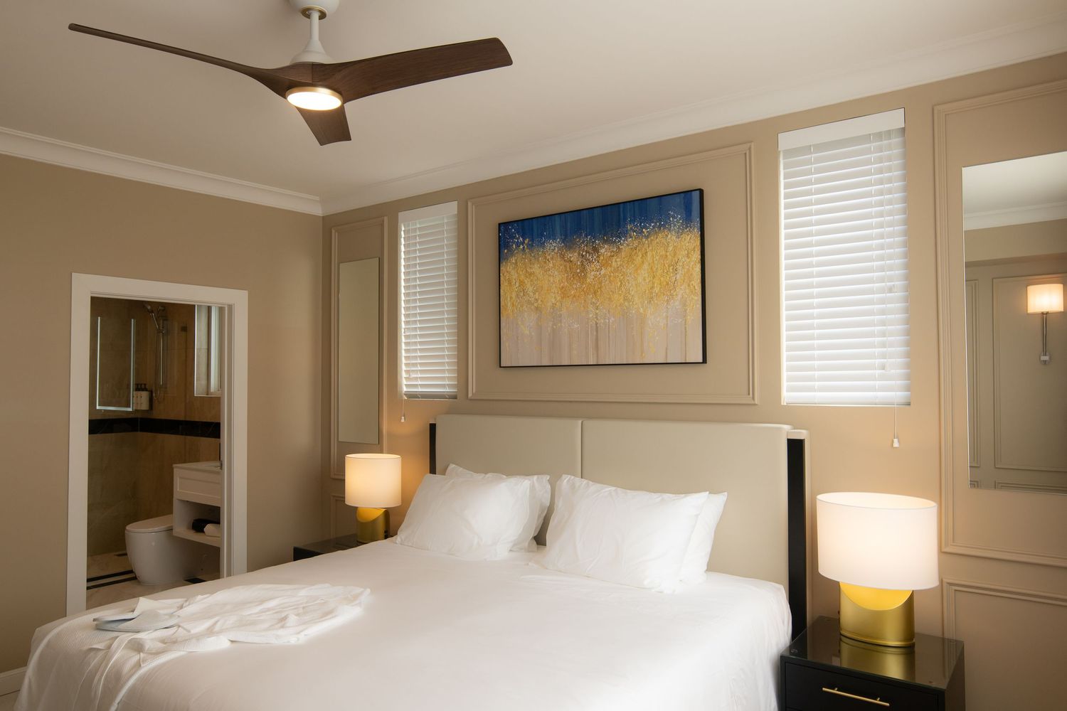 Elegant hotel bedroom with white bedding, ceiling fan, bedside lamps, abstract wall art, and an ensuite bathroom.