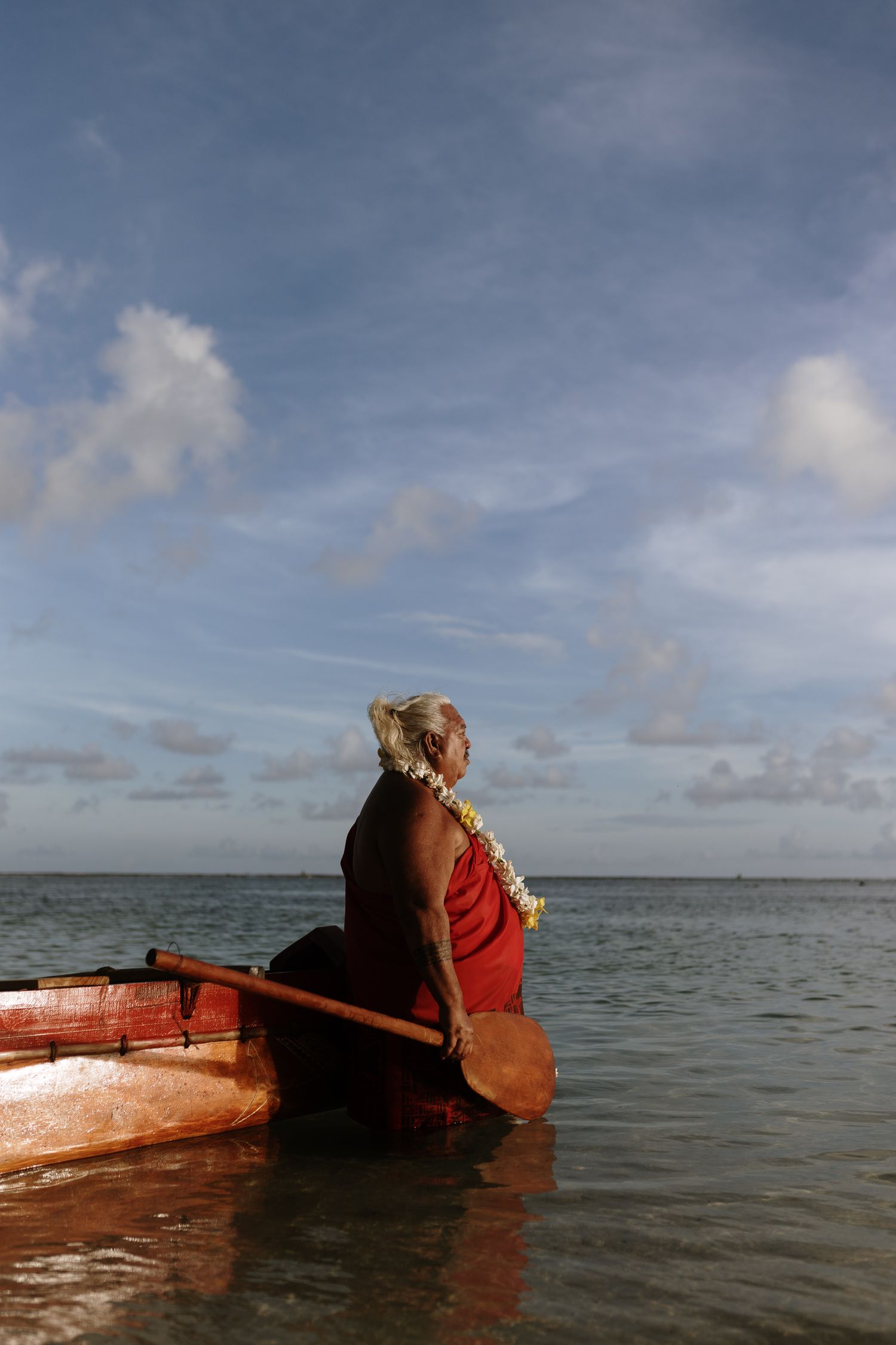 Elderly Pacific Islander in red garment and floral lei sits in wooden canoe, paddling on calm tropical ocean.