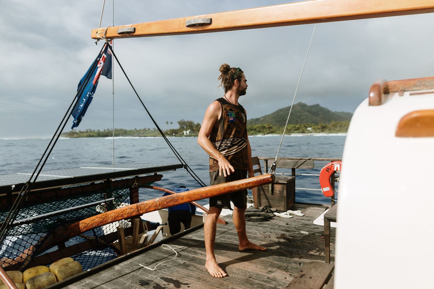 Barefoot sailor stands on wooden deck of traditional sailboat with ocean and tropical island in background.