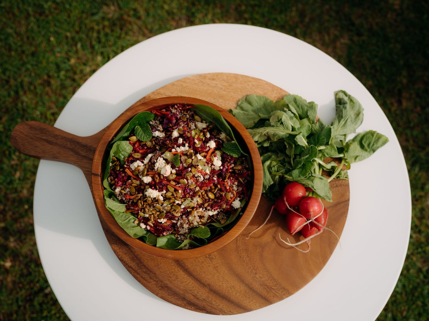 Wooden bowl filled with quinoa beet salad topped with feta, served on a cutting board with fresh radishes and greens.