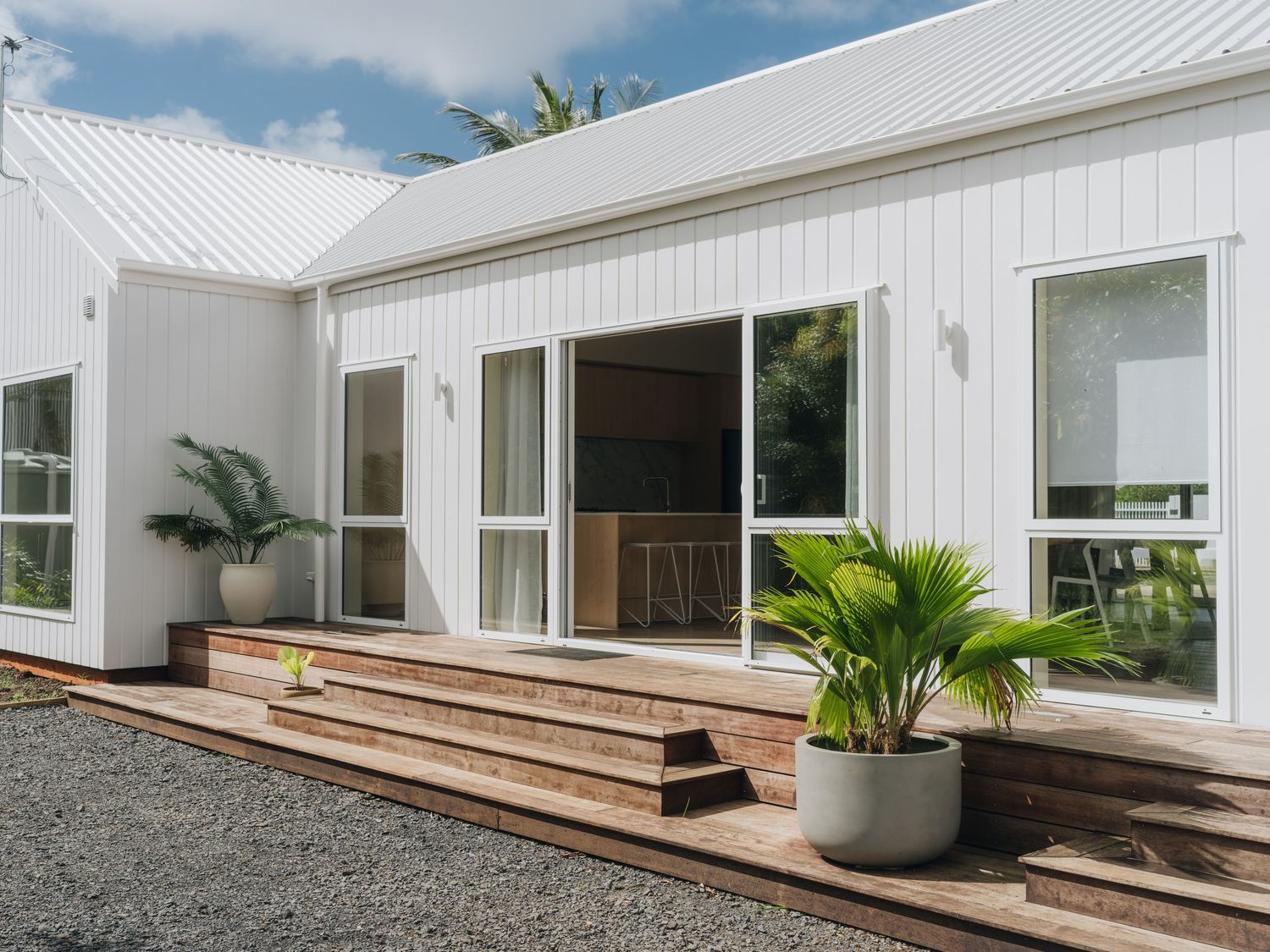 White modern home exterior with wooden deck steps, sliding glass doors, and tropical potted plants surrounded by gravel.