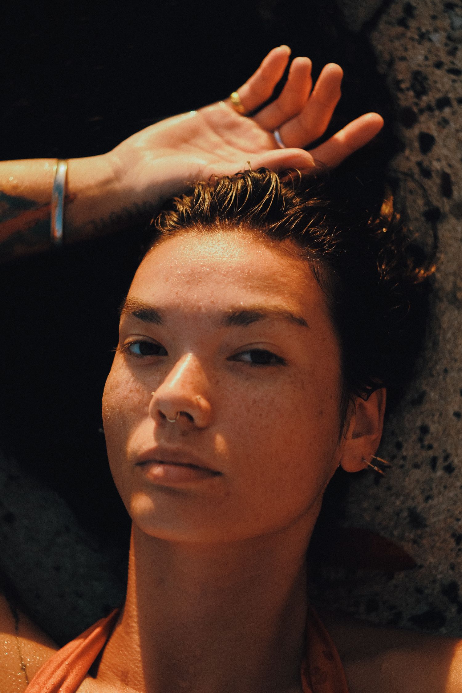 Close-up portrait of a young woman with freckles, wet hair, and a nose ring, resting against a dark rock surface.