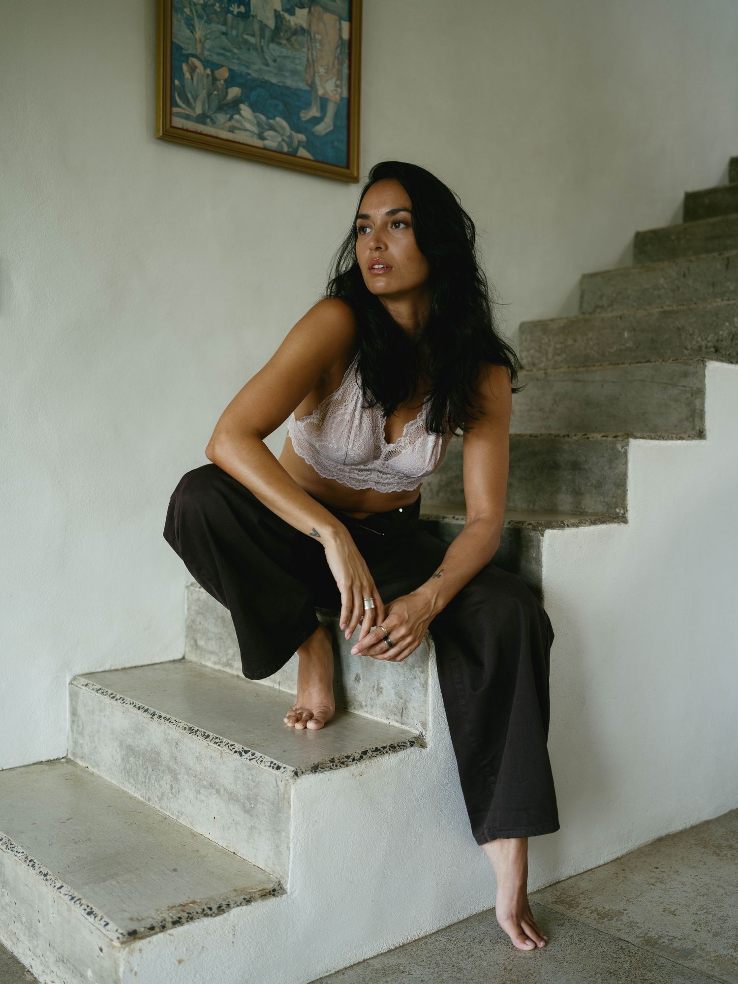Woman in black wide-leg pants and white crop top sitting barefoot on concrete stairs near a framed painting.