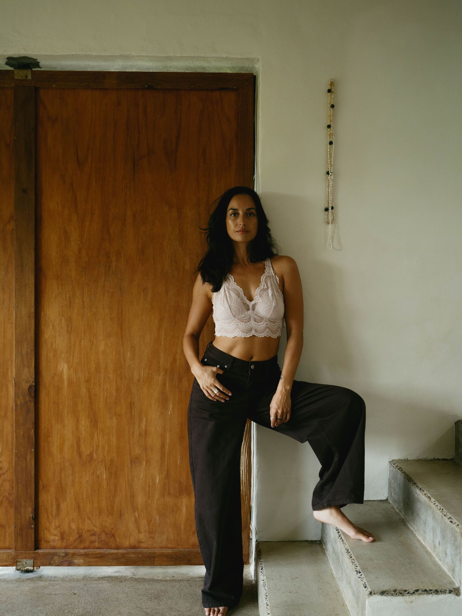 Woman in white lace crop top and black wide-leg pants posing barefoot on steps near a wooden door.