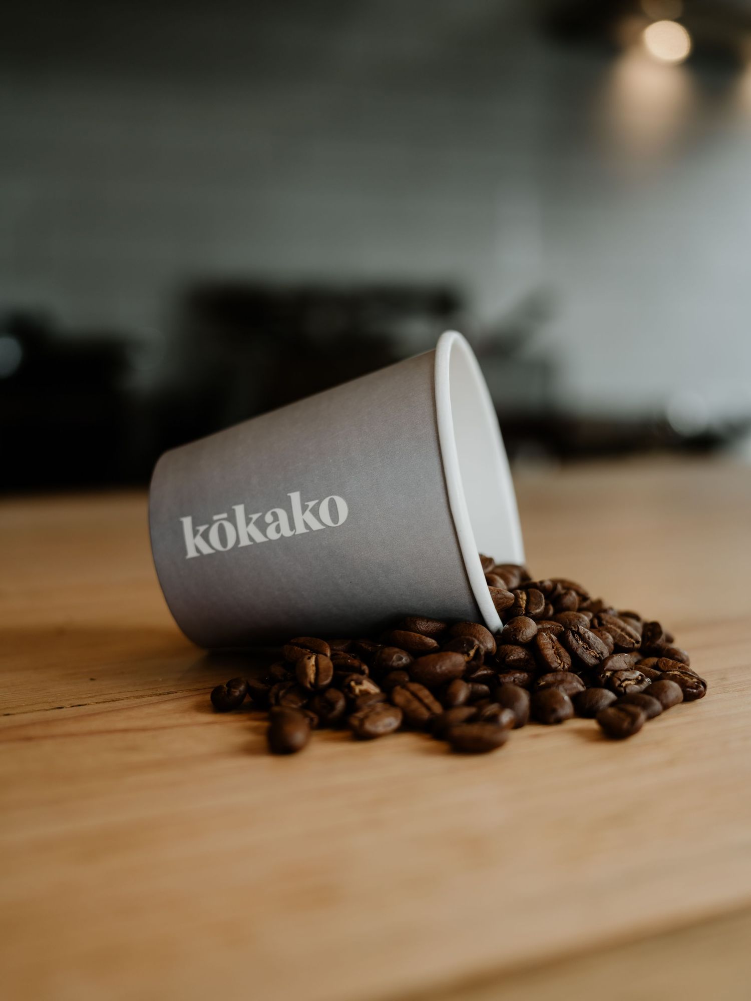 A Kokako branded grey paper cup tipped on its side spilling roasted coffee beans on a wooden surface.