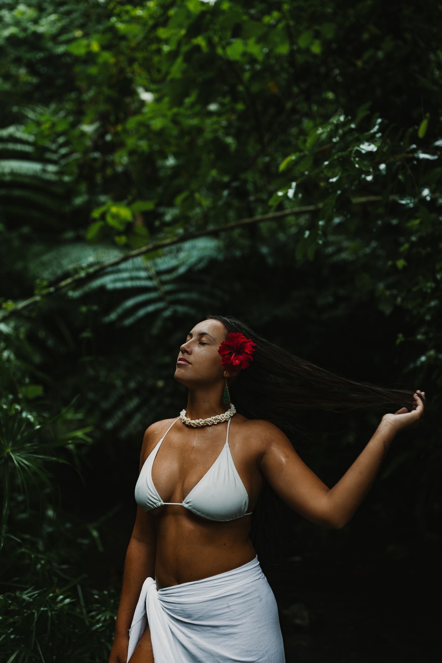 Woman in white bikini top and sarong with red flower in hair poses in lush tropical jungle setting.