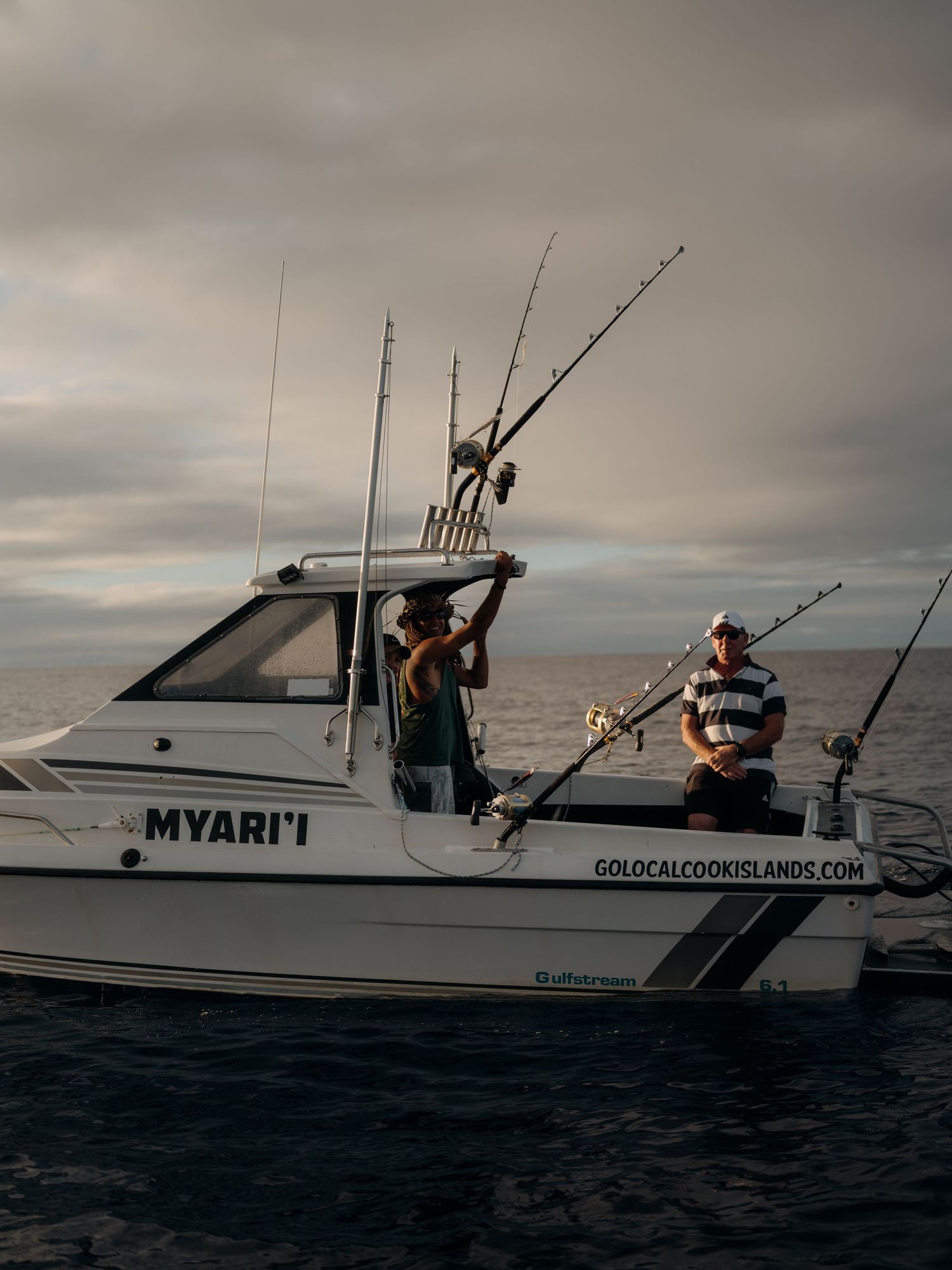 Two anglers fishing from the MYARI'I charter boat on calm ocean waters, advertised by GoLocalCookIslands.com.