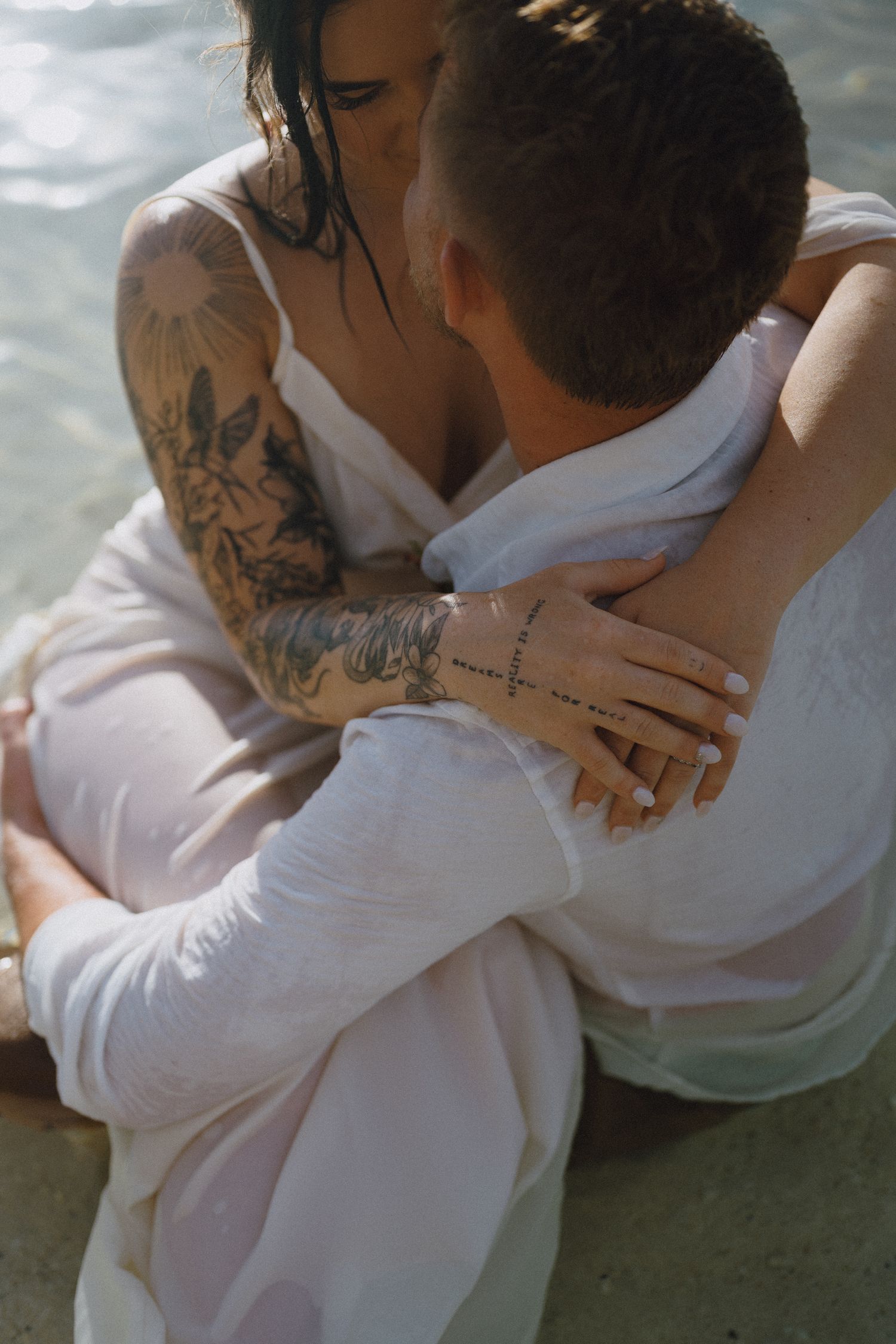 Couple embracing intimately near water, woman with floral tattoos in white dress hugging man in white shirt.