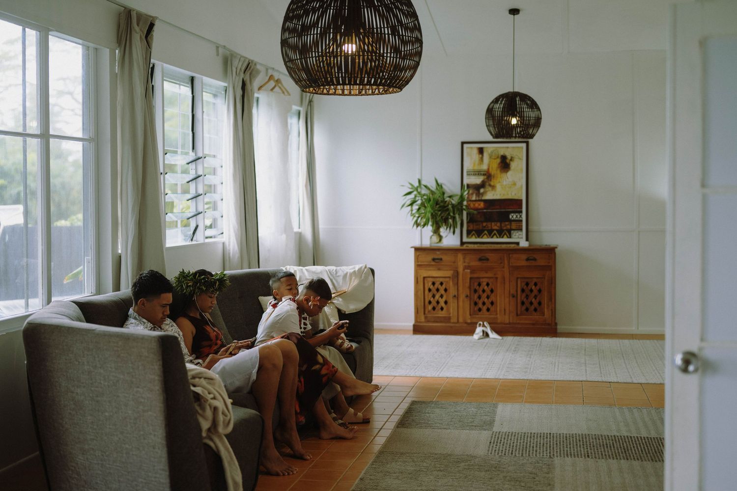 Family relaxing together on a gray sofa in a modern living room with wicker pendant lights and wooden sideboard.