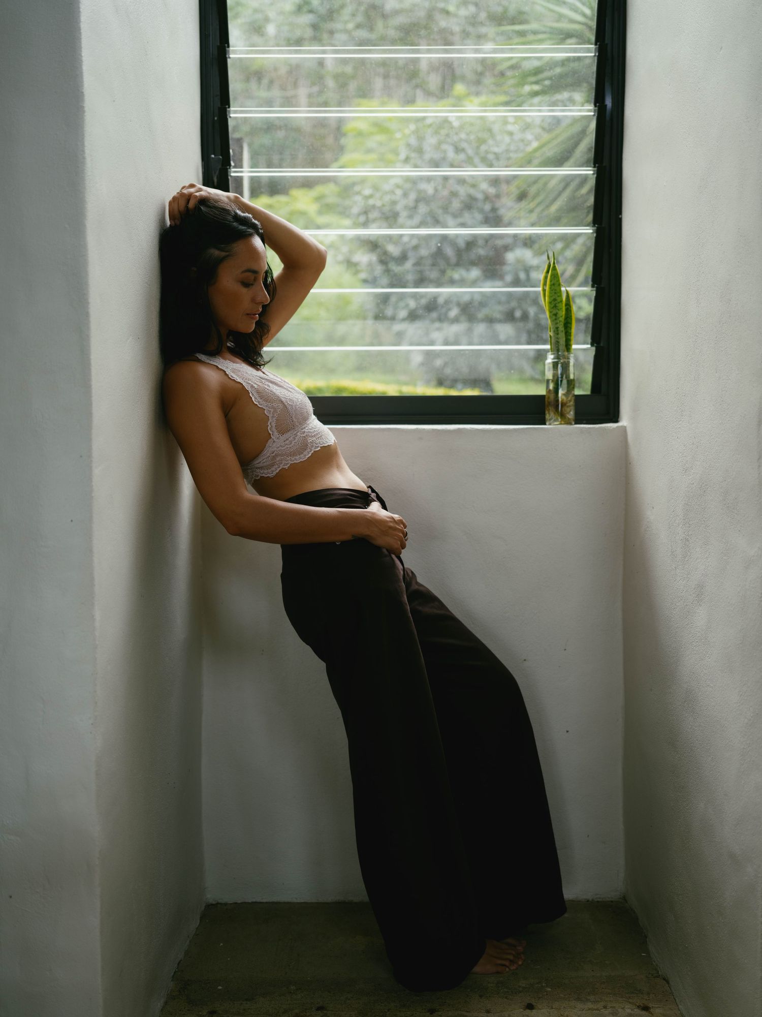 Woman in white crop top and black wide-leg pants leaning against white wall near window with greenery.