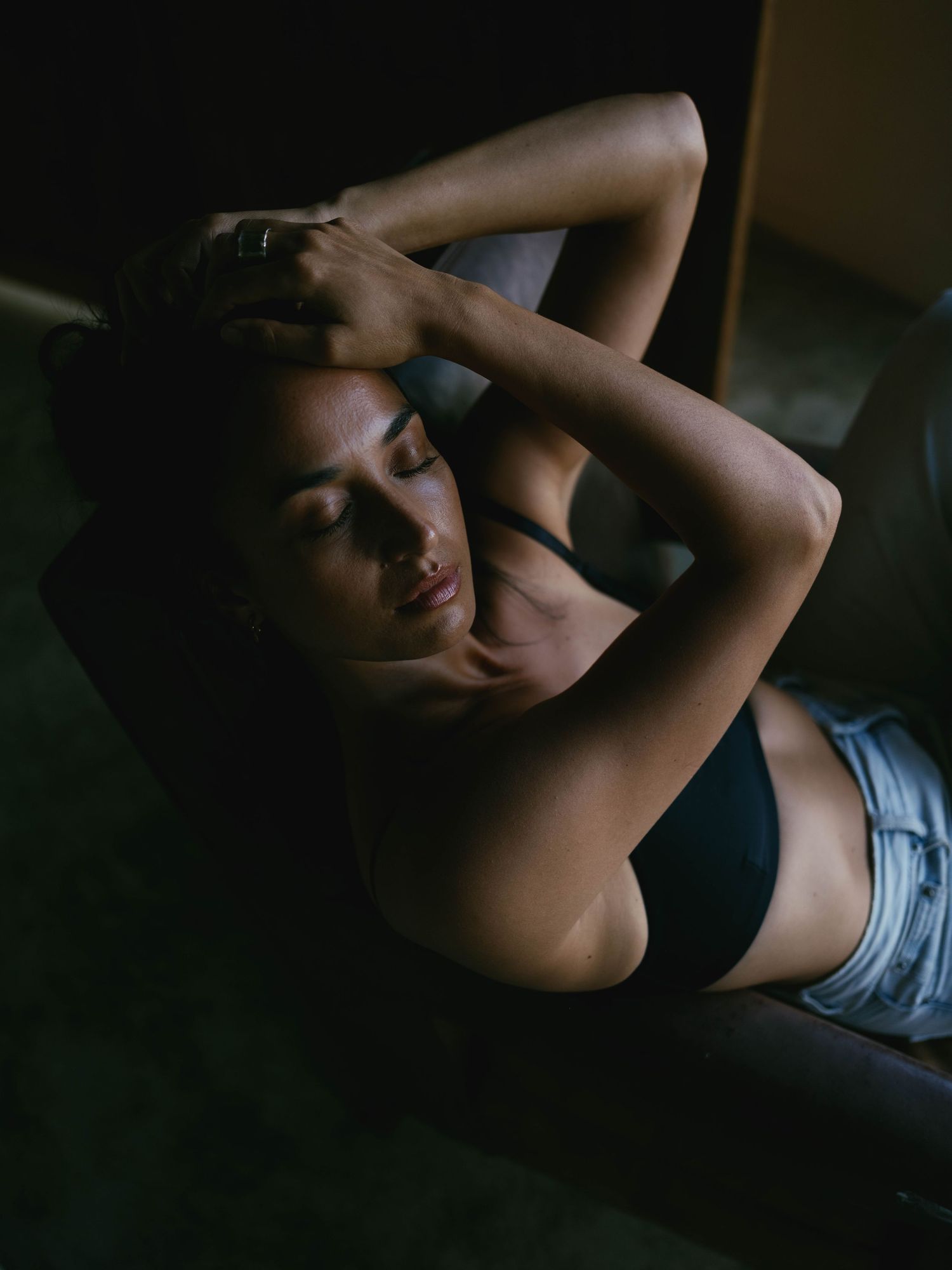 A woman in a dark bra and denim shorts reclines on a sofa with arms raised, eyes closed, in moody lighting.