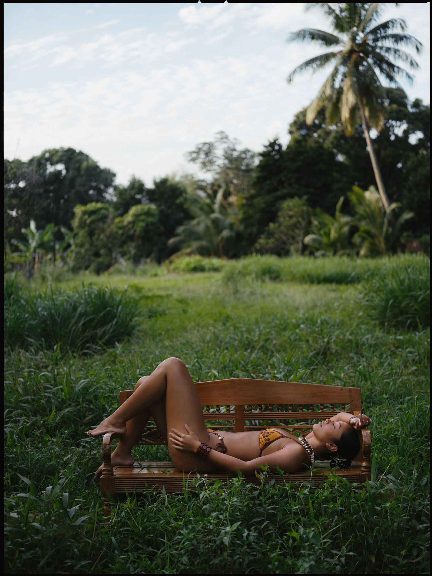 Woman in bikini reclining on wooden bench surrounded by lush tropical greenery and palm trees.