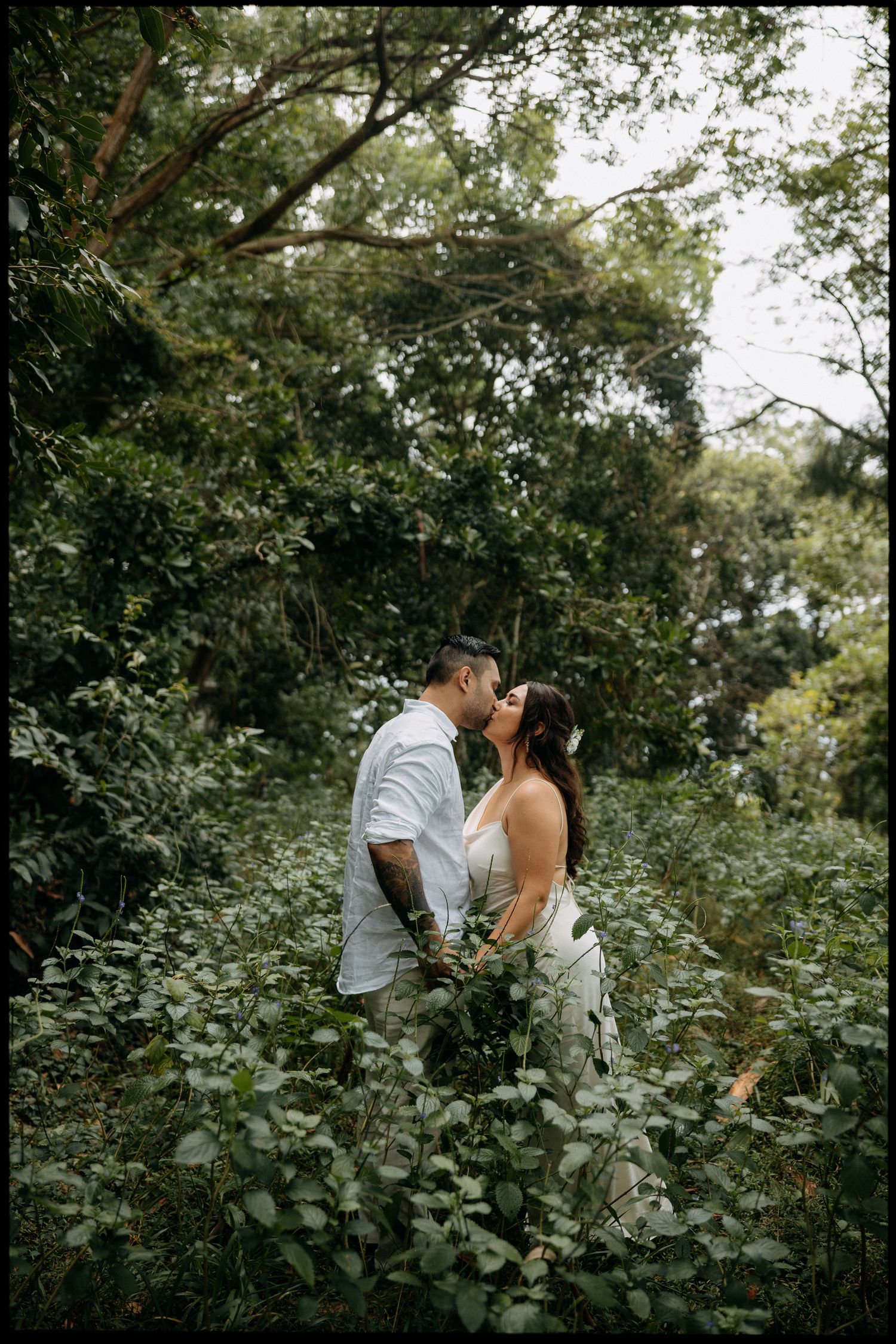 A couple shares a romantic kiss surrounded by lush green foliage in a forest, dressed in wedding attire.
