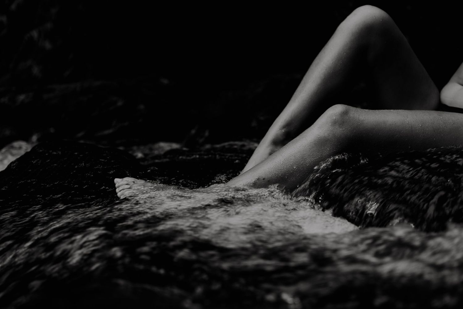 Black and white photo of bare legs resting on rocks beside flowing water in a serene outdoor setting.