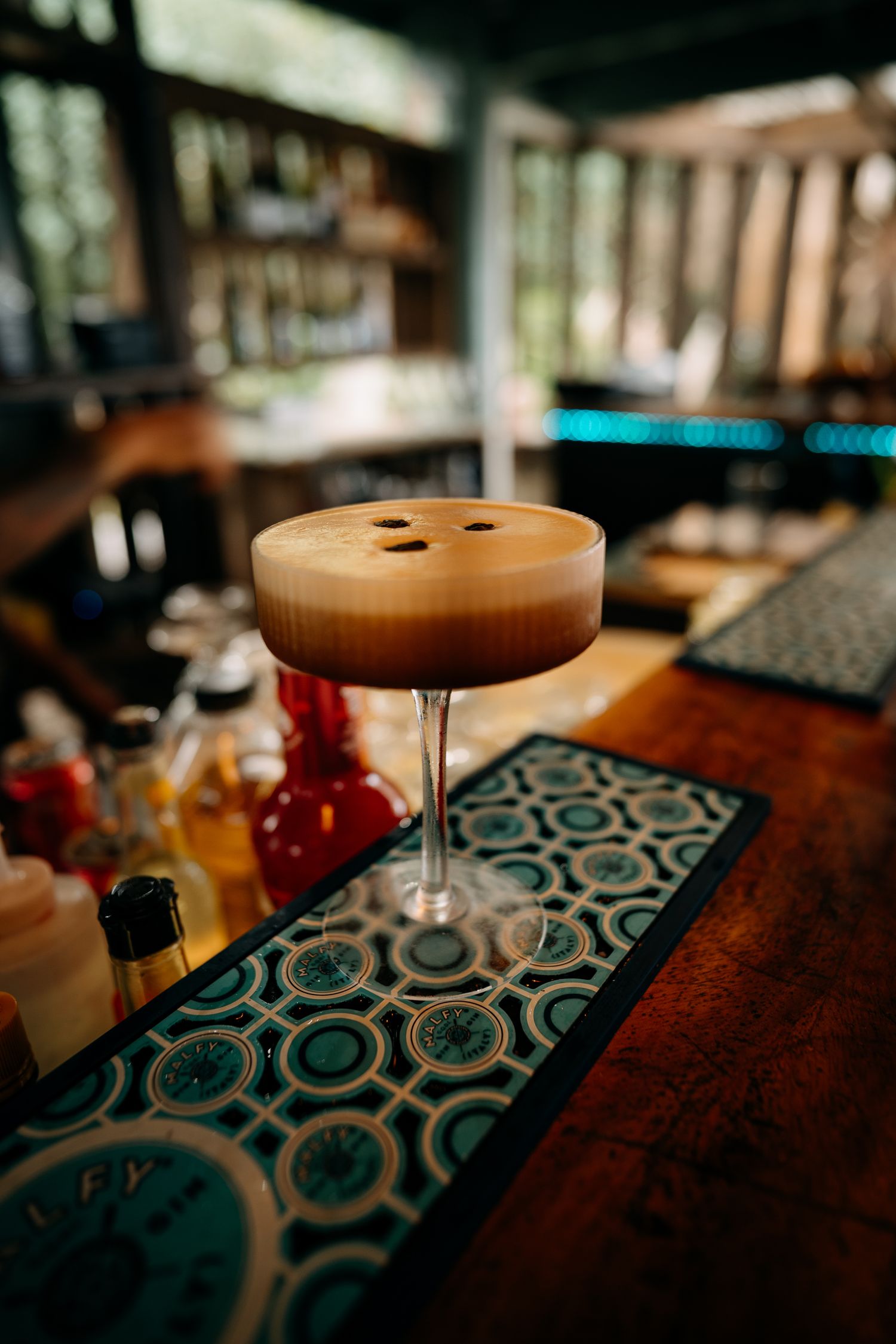 Espresso martini cocktail with frothy top and coffee beans served in coupe glass on teal bar mat.
