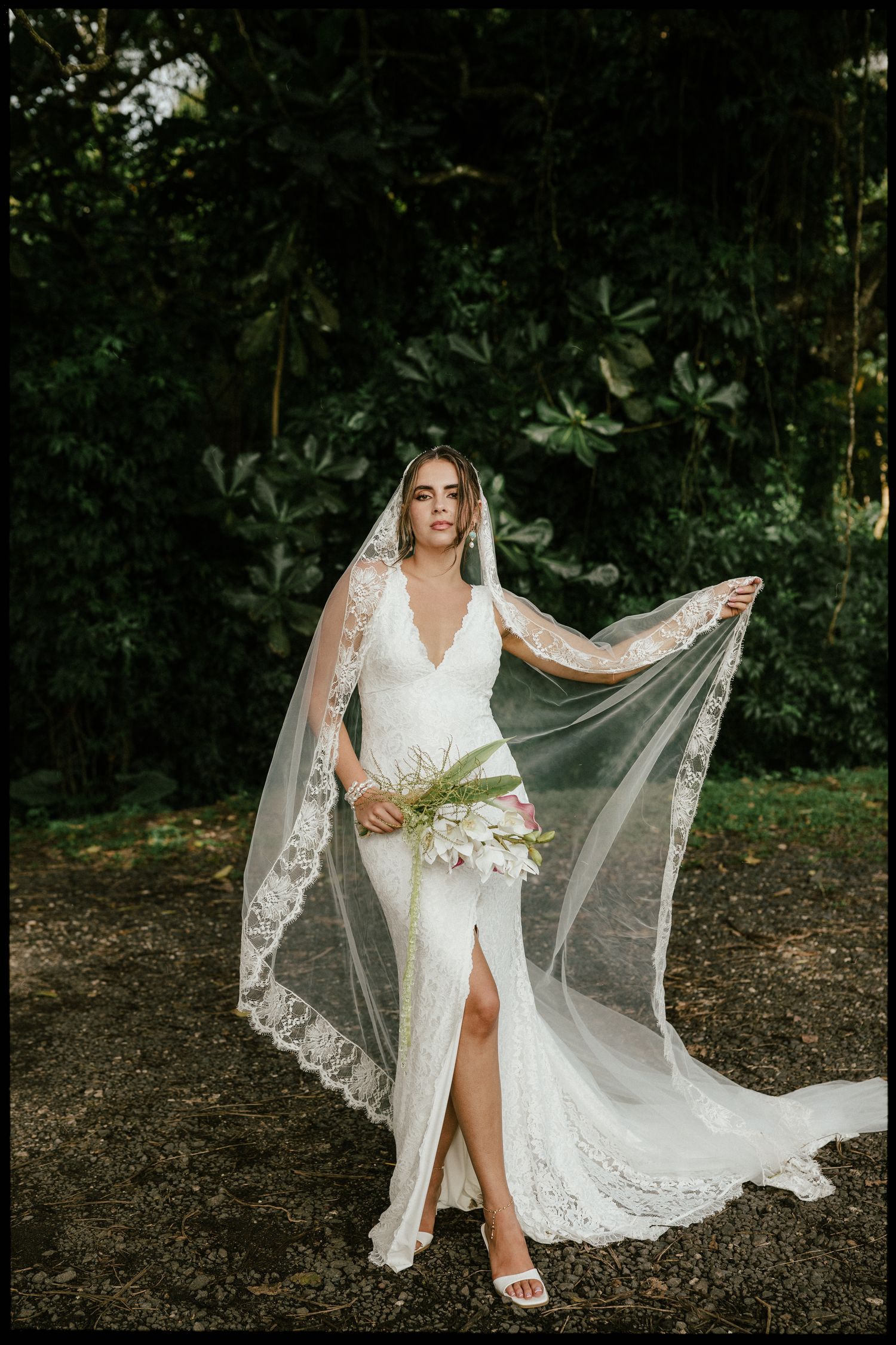 Bride in white lace wedding gown with flowing veil holding bouquet, standing outdoors among lush green tropical foliage.