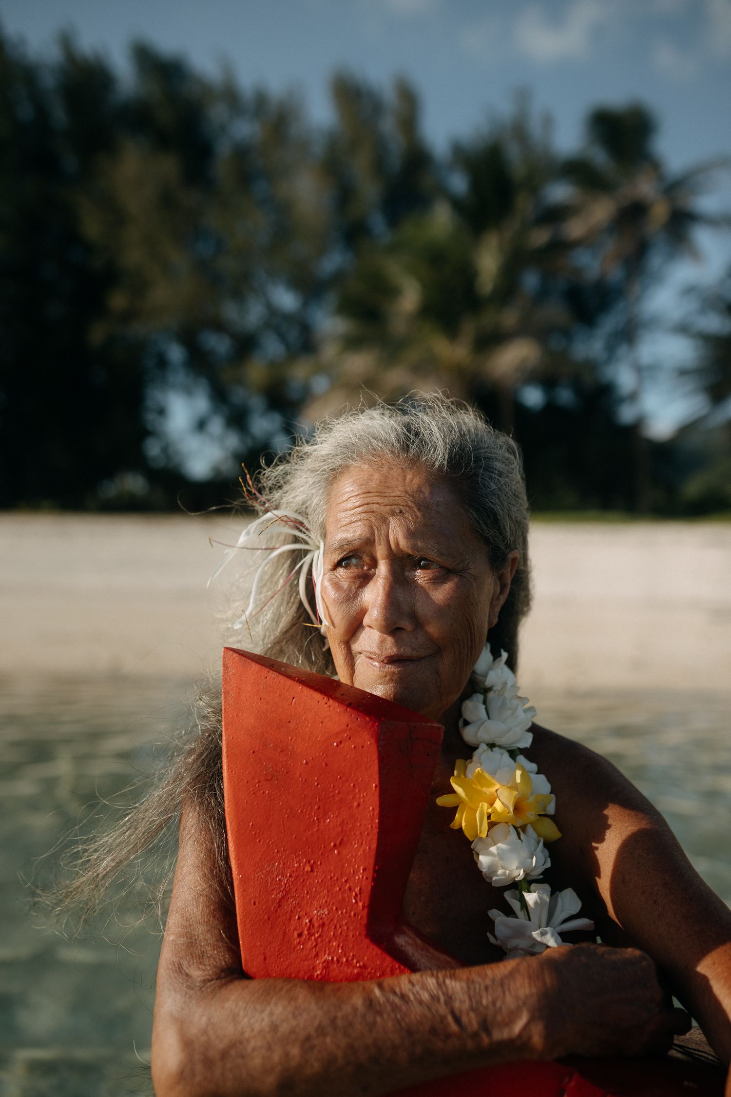 Elderly Pacific Islander woman with gray hair and floral lei holds red board on tropical beach.