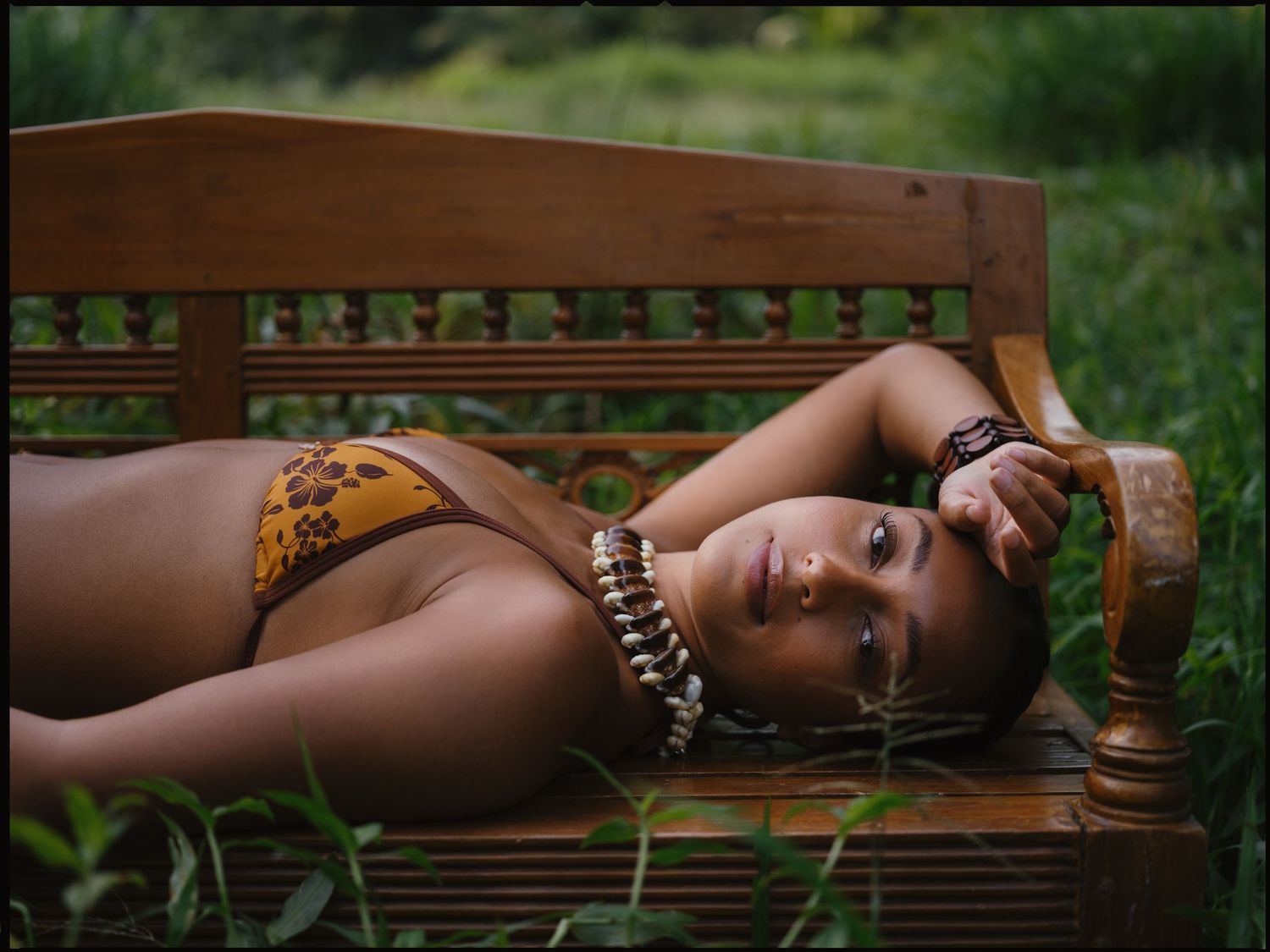 Woman in bikini lying on wooden bench surrounded by lush green foliage outdoors.