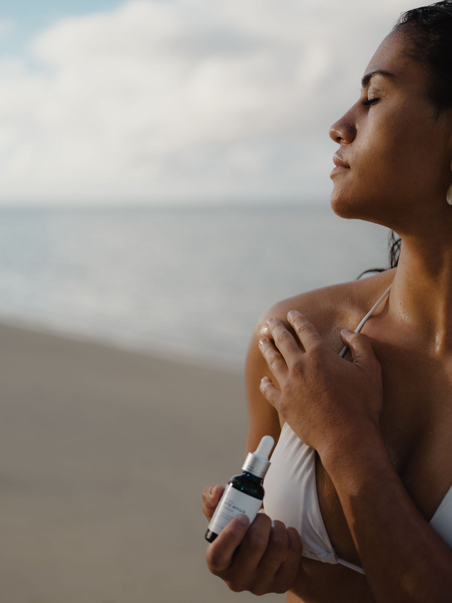 Woman applying skincare serum on her shoulder at the beach, eyes closed, holding a dropper bottle, with ocean in background.