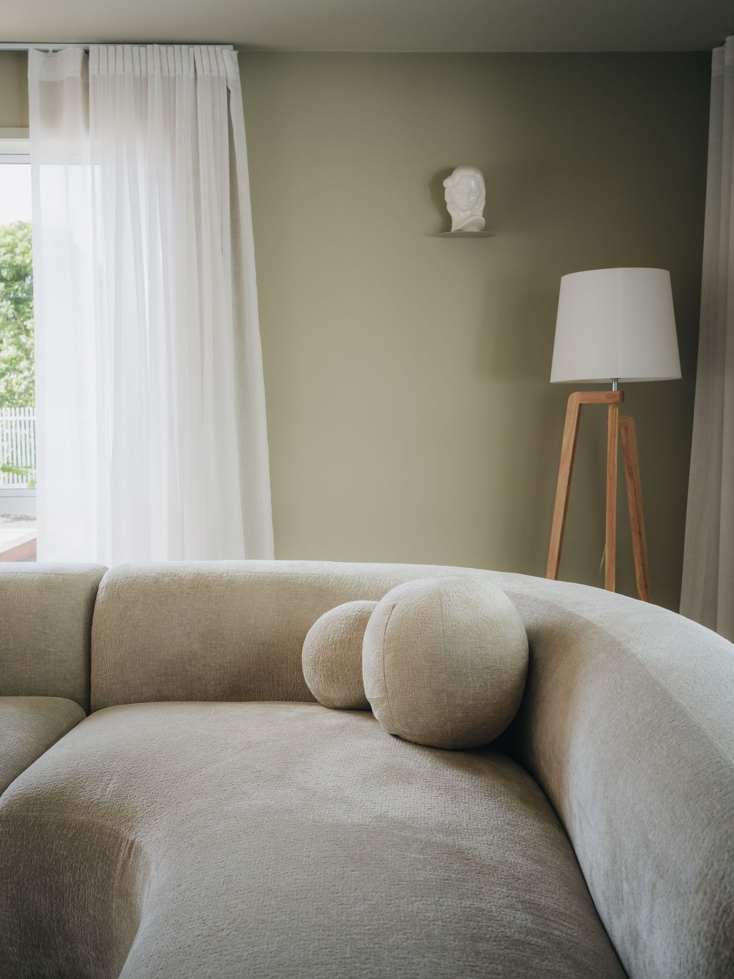 Minimalist living room with beige curved sofa, round pillows, tripod floor lamp, and sage green walls with sheer curtains.