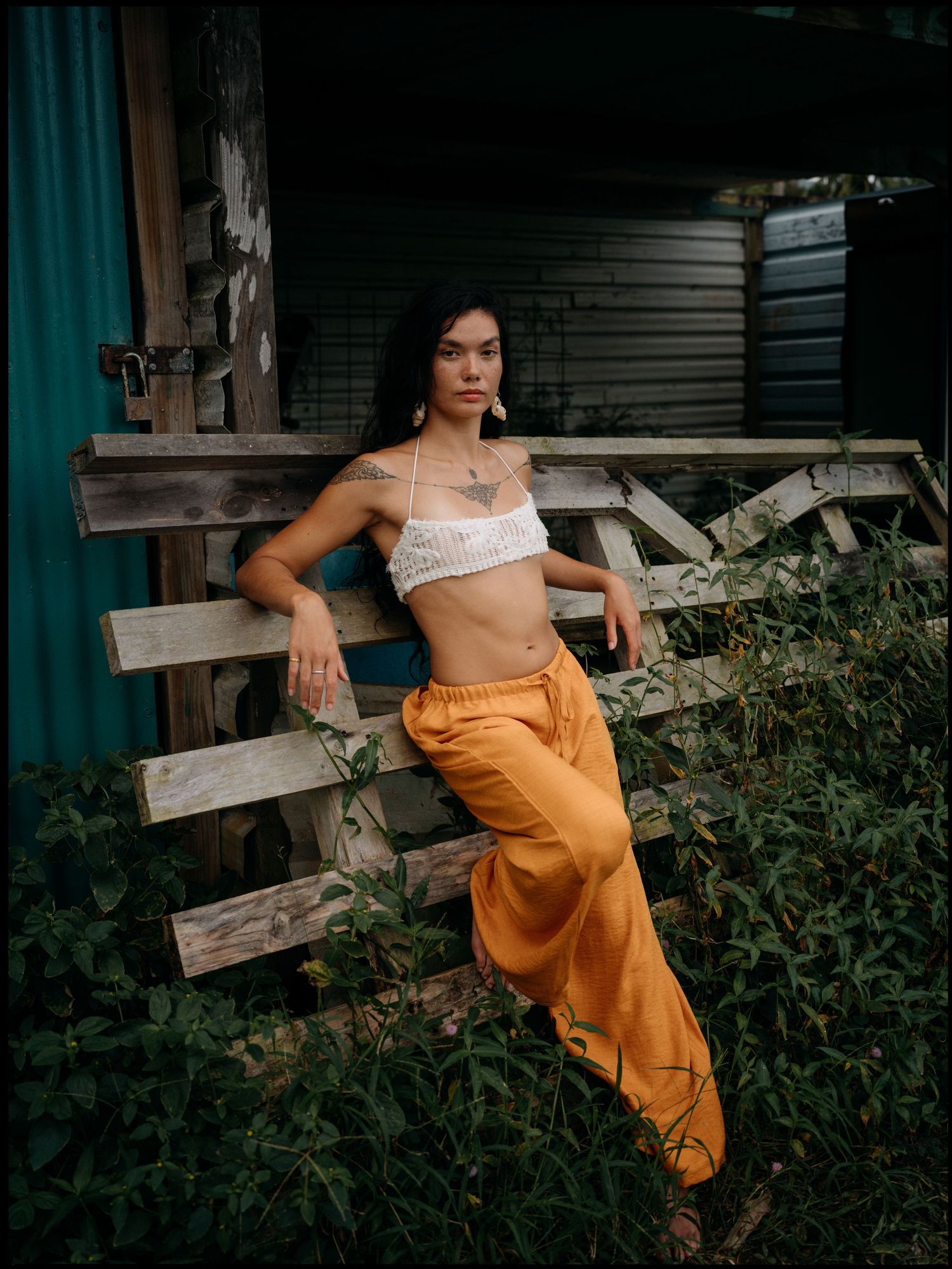 Woman in white crop top and orange wide-leg pants leaning on rustic wooden fence surrounded by lush greenery.