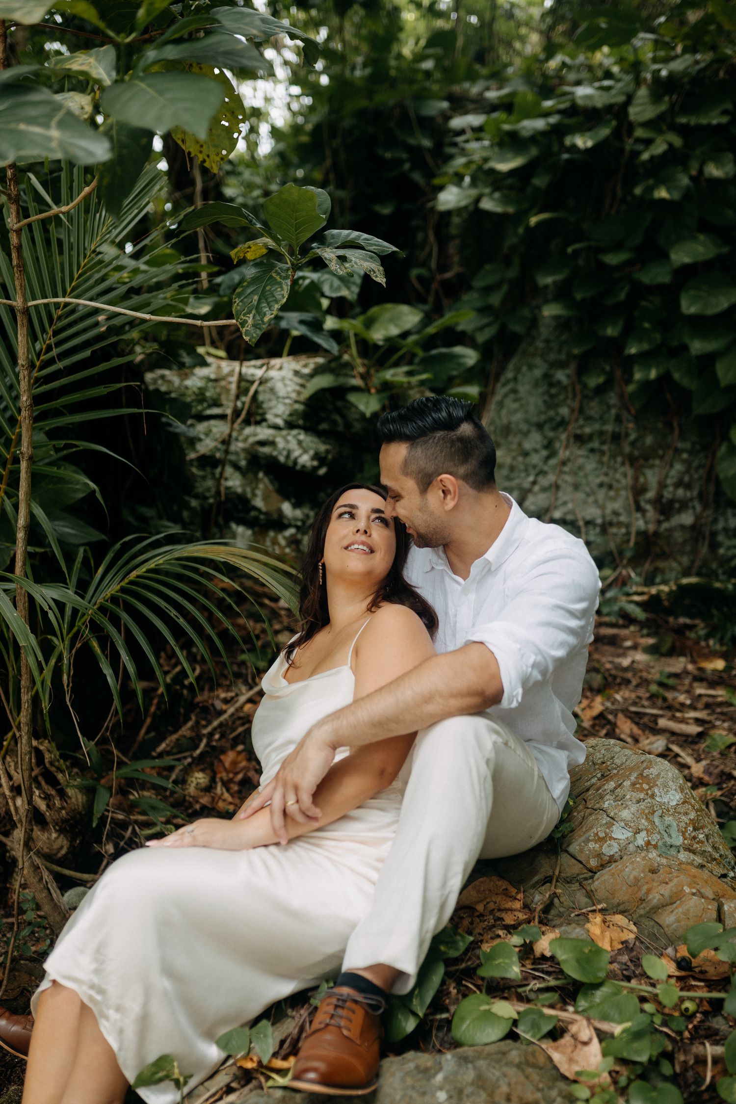 A couple in white attire sits romantically among lush tropical greenery, the man gently kissing the woman's cheek.