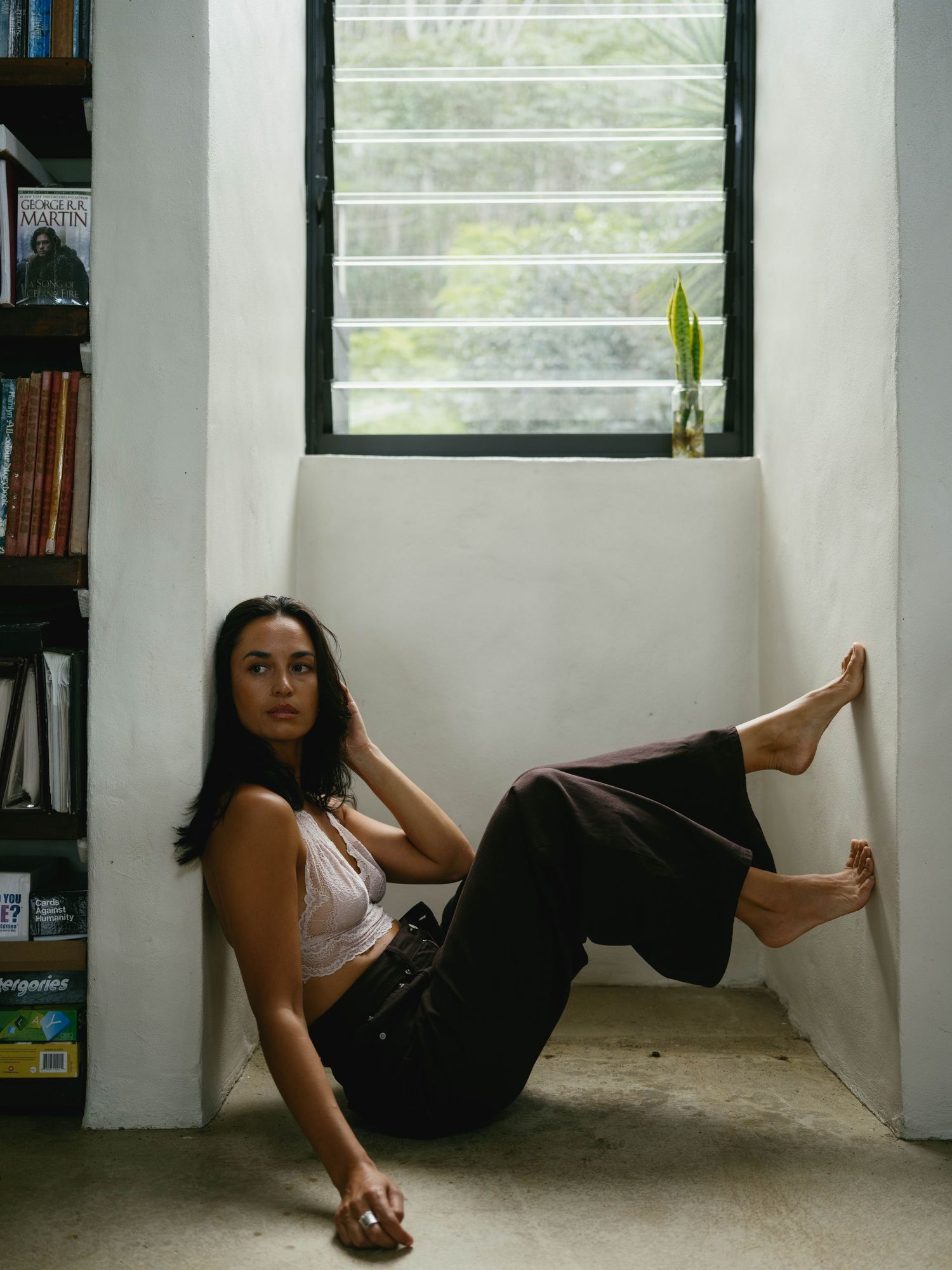 Woman in white top and dark pants sitting relaxed against white wall near window and bookshelf.