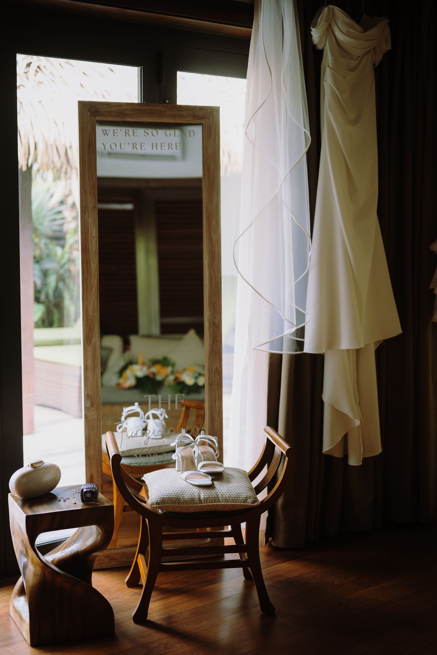 Elegant wedding dress hanging near a mirror with bridal shoes on a wooden chair in a tropical villa setting.