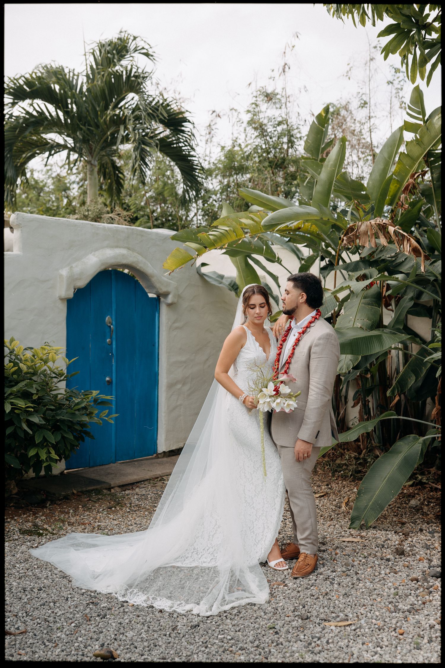 Bride and groom pose near a white stucco wall with blue door, surrounded by tropical palms and lush greenery.