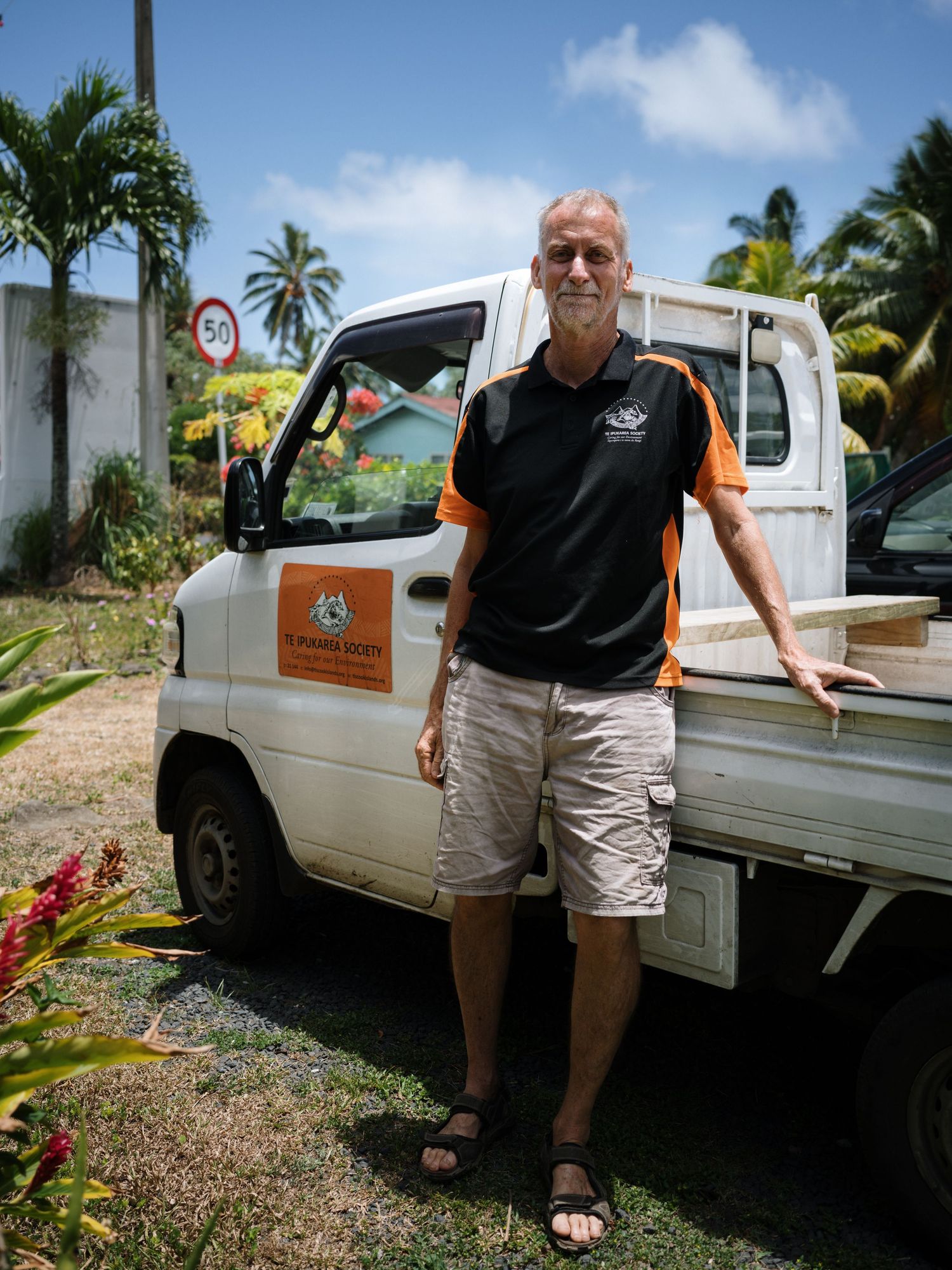 Man in black and orange polo shirt stands beside a white utility truck in a tropical setting.