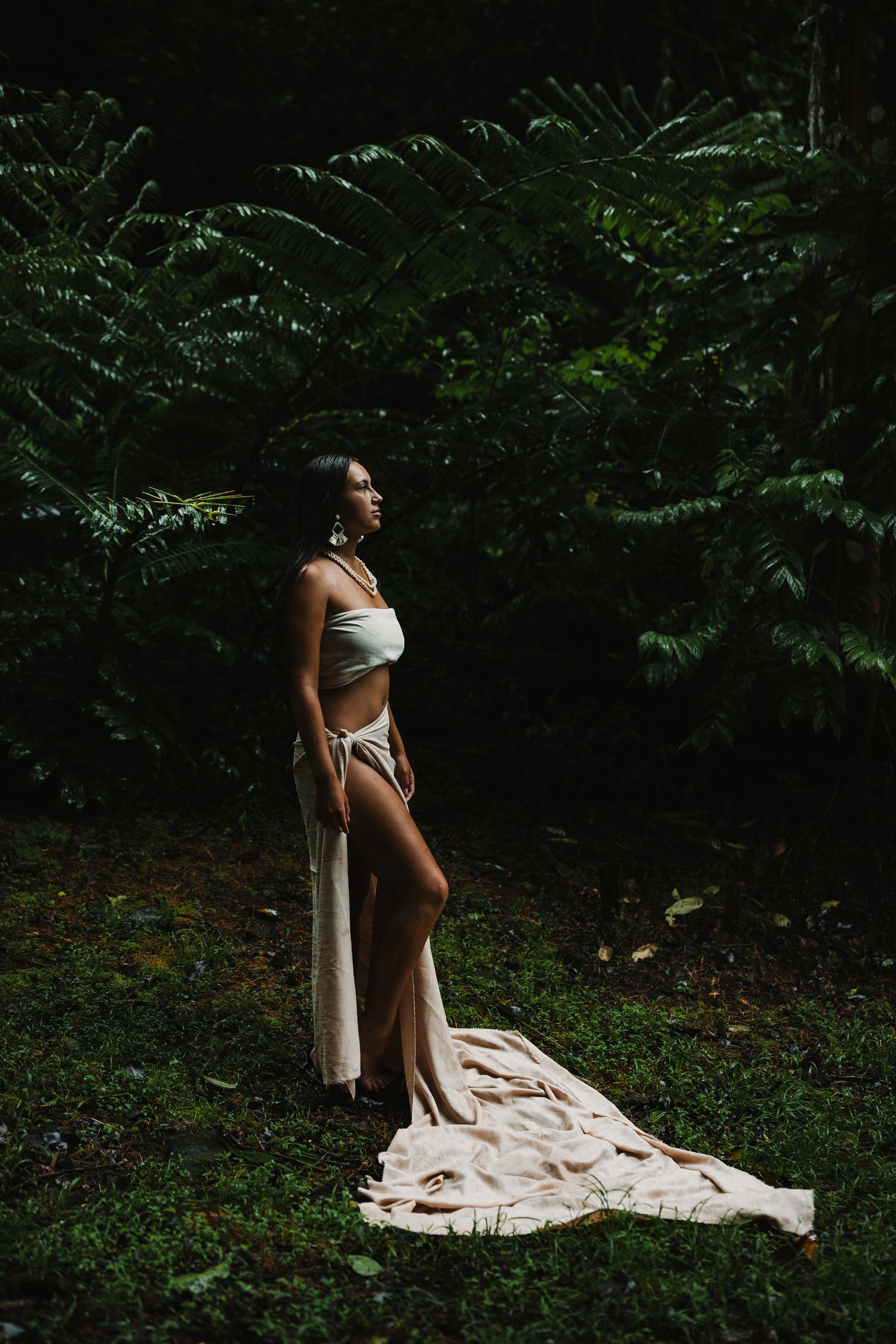 Woman in white bikini and flowing skirt poses gracefully in lush tropical forest setting.