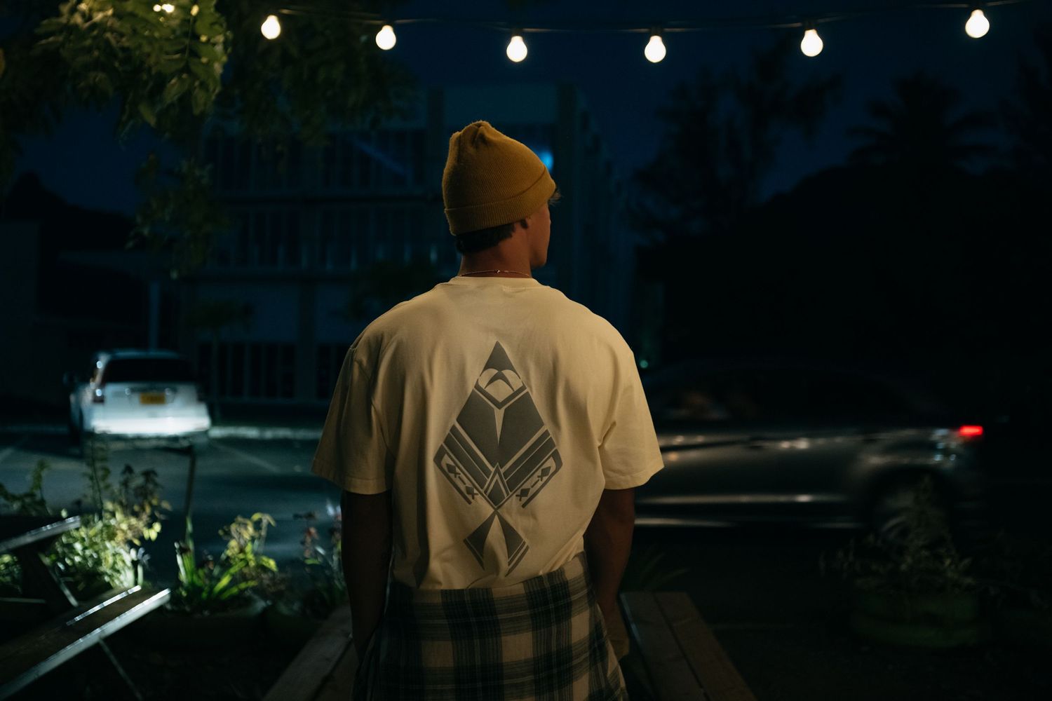 Man in white graphic tee and plaid shorts stands on a deck at night under string lights.