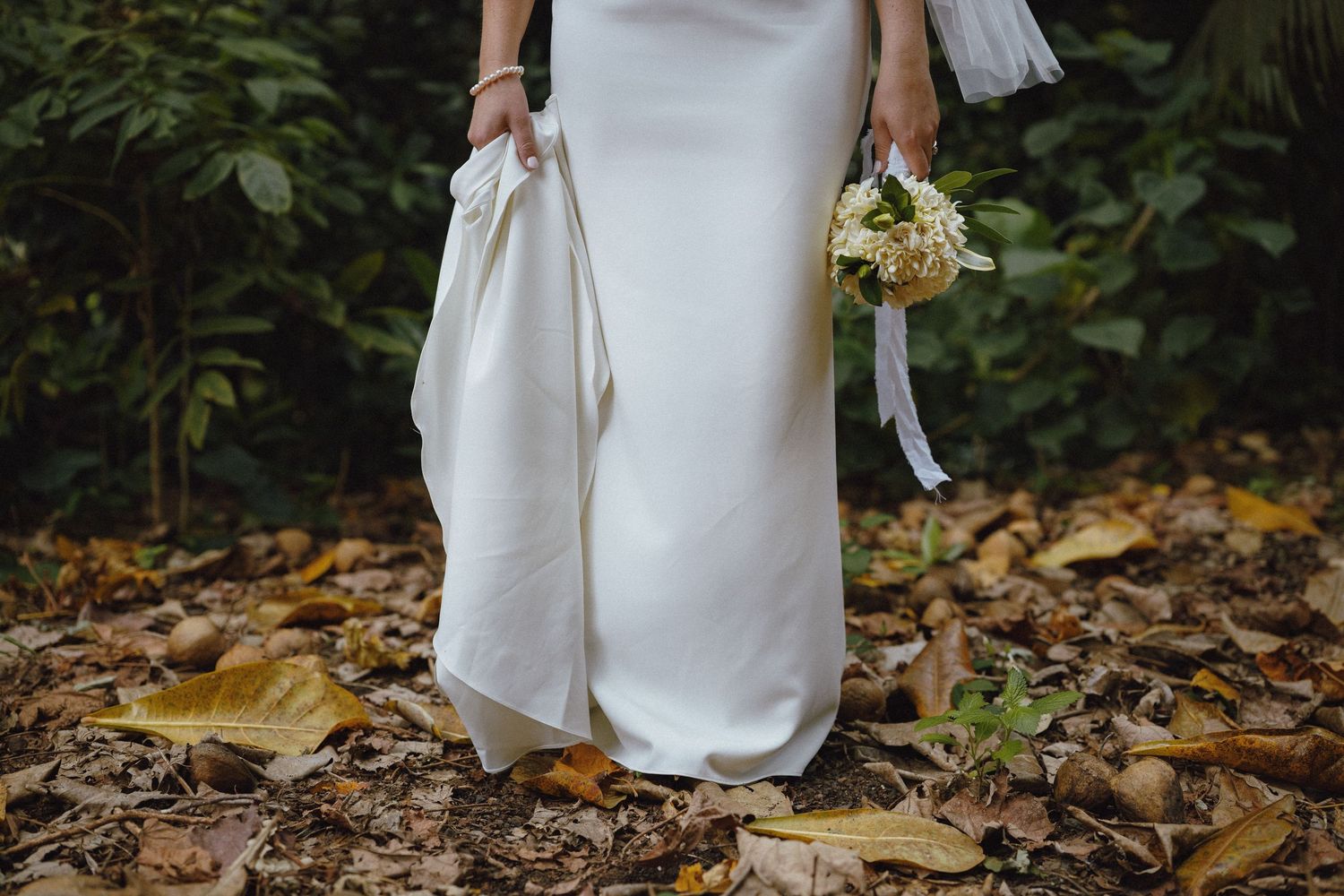 Bride in white satin gown holding ivory floral bouquet, standing on autumn leaves in a lush garden setting.