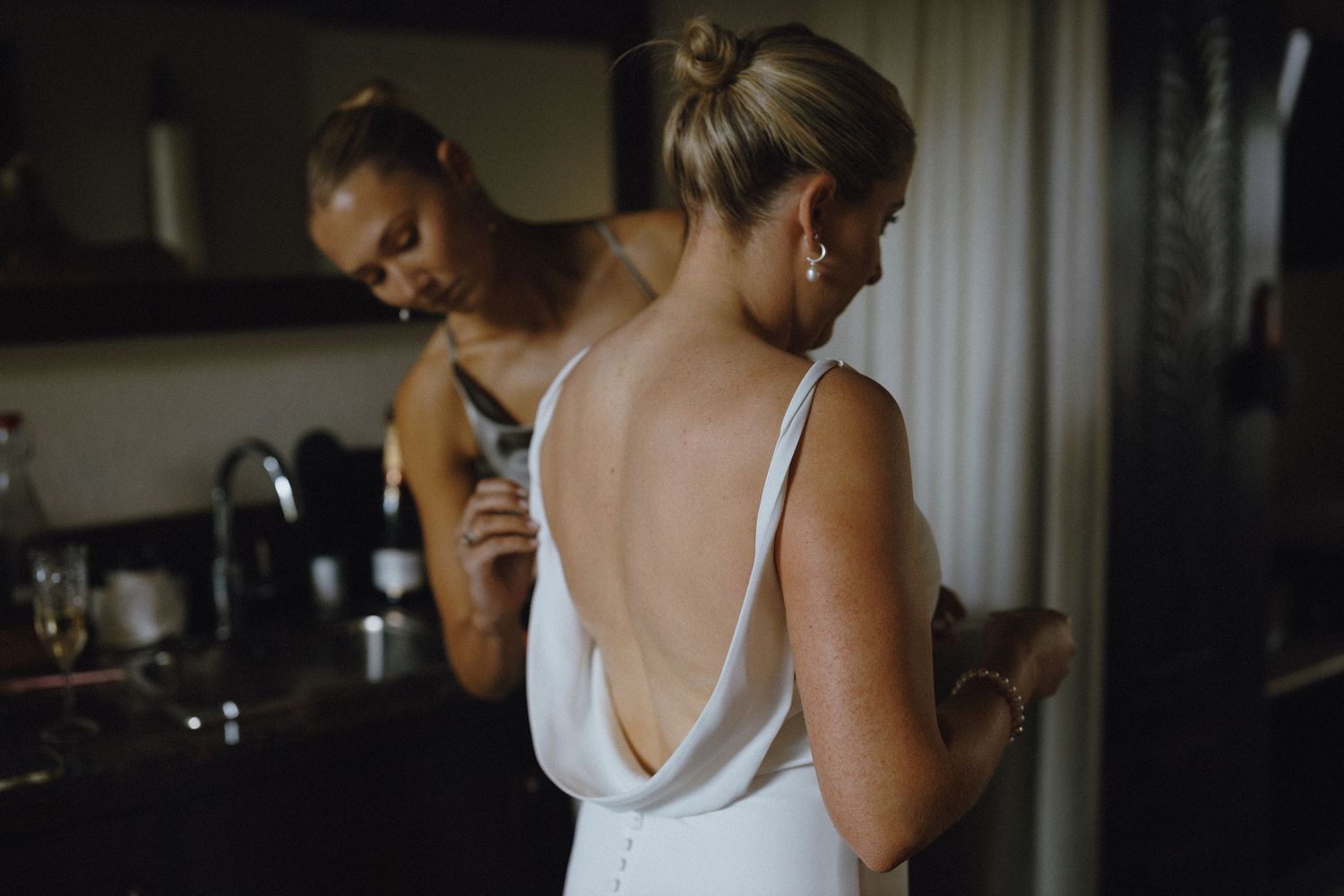 Bride in elegant backless white wedding gown being assisted by bridesmaid, reflected in mirror during bridal preparations.