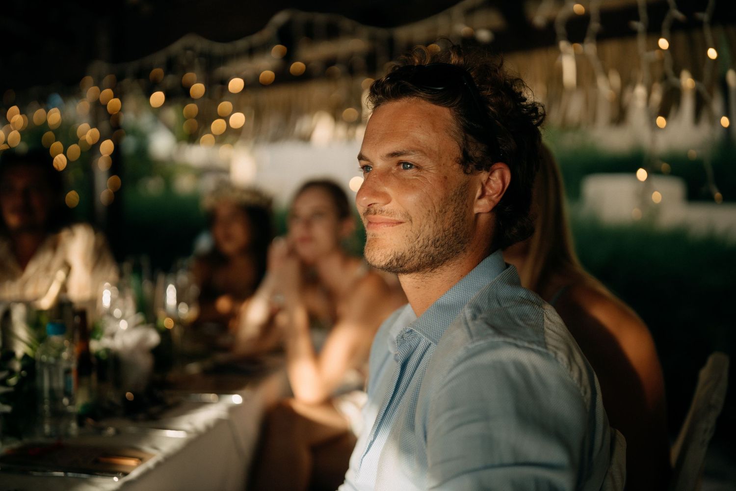 A smiling man in a light blue shirt sits at an outdoor dinner party with bokeh string lights in the background.