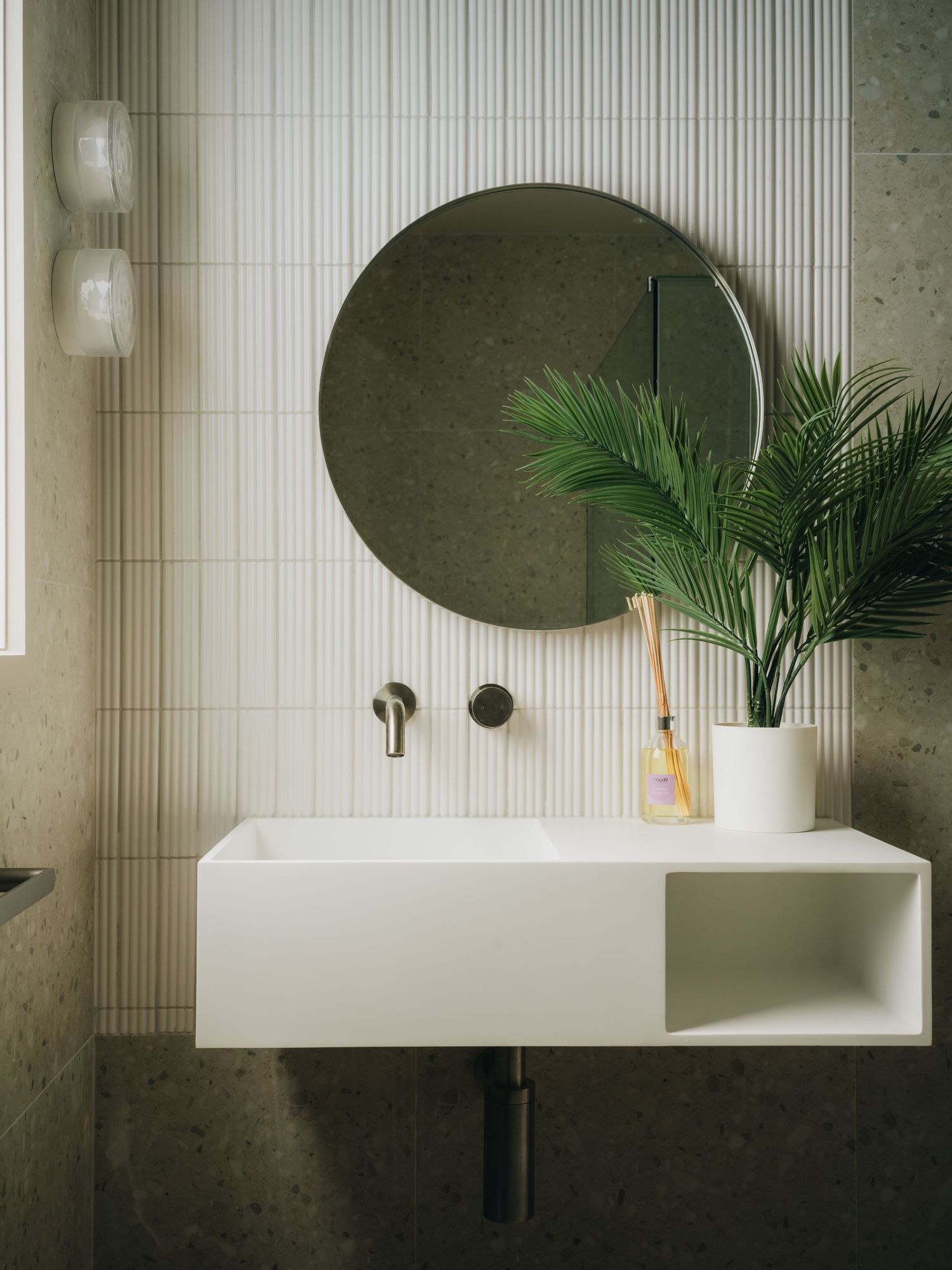 Modern bathroom with round mirror, white floating sink, green plant, and vertical tile wall.