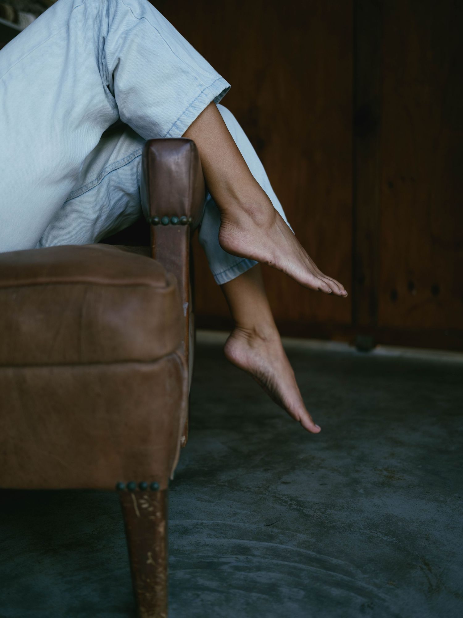 Bare feet dangling over the arm of a vintage brown leather chair, wearing light blue denim shorts.