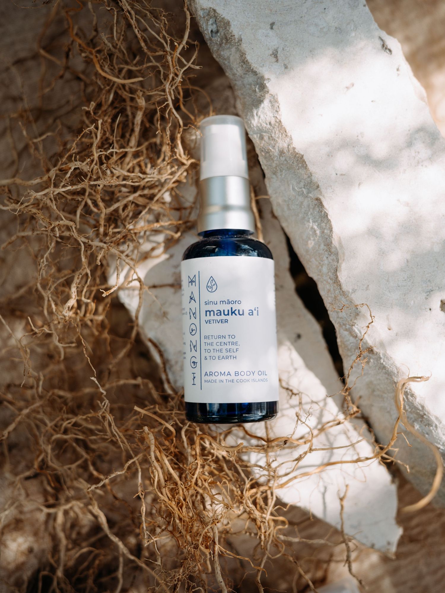 Mauku a'i aroma body oil spray bottle resting on natural stone and dried grass in an earthy outdoor setting.