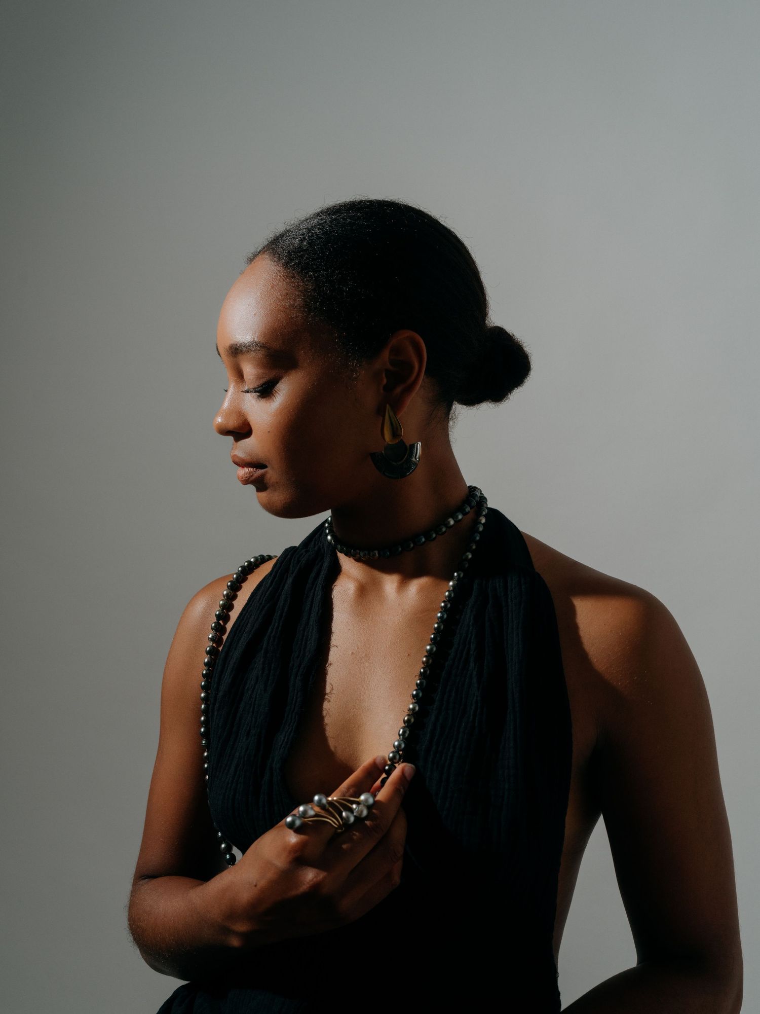 Woman in black halter dress with beaded necklace, hoop earrings, and statement ring poses gracefully in studio.