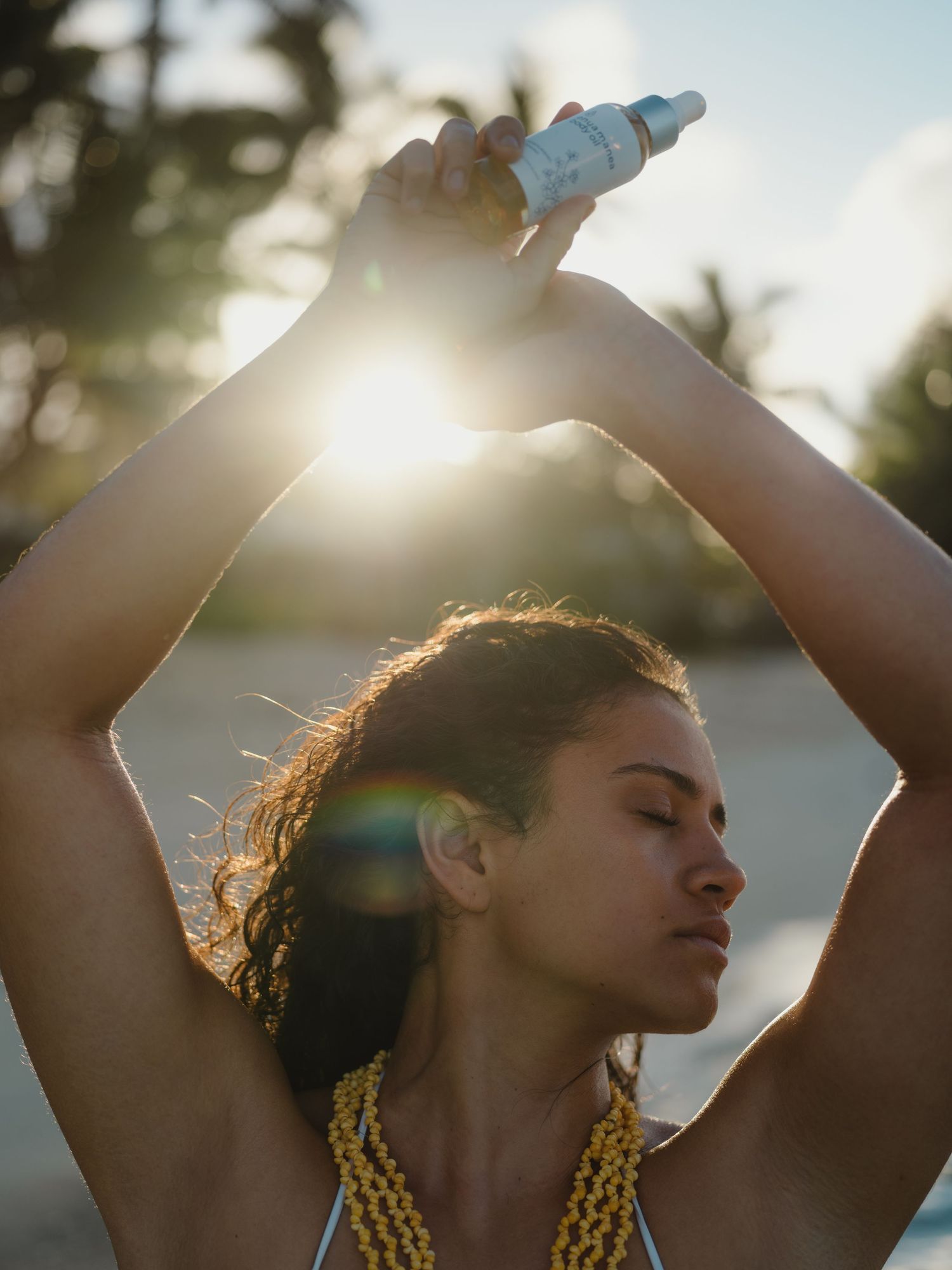Woman applying sunscreen spray at sunset on a beach, wearing a yellow bikini top with sun flare behind her.