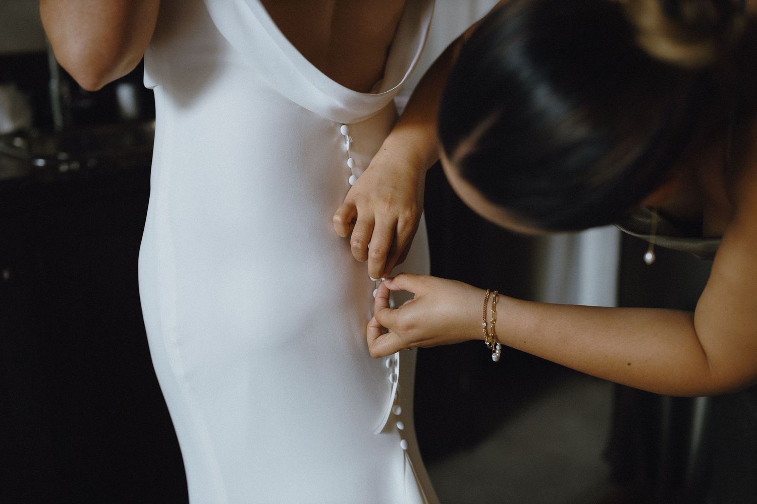 A bridesmaid carefully fastening pearl buttons on the back of a white wedding dress.