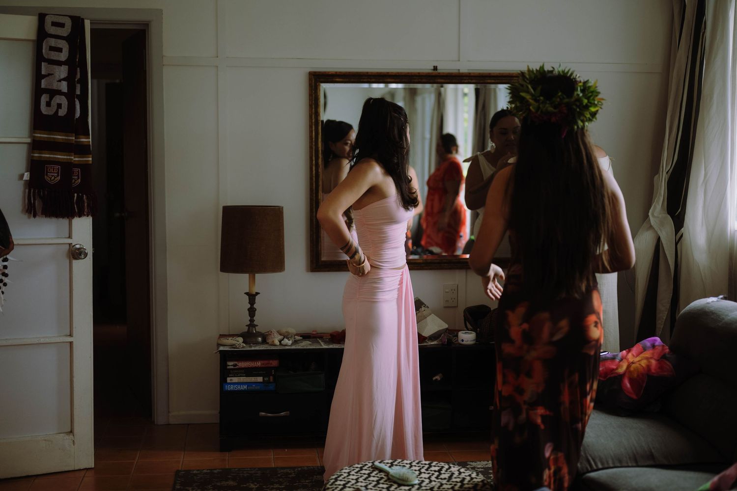 Two bridesmaids in pink dresses prepare for a wedding, reflected in a vintage mirror in a warmly lit room.