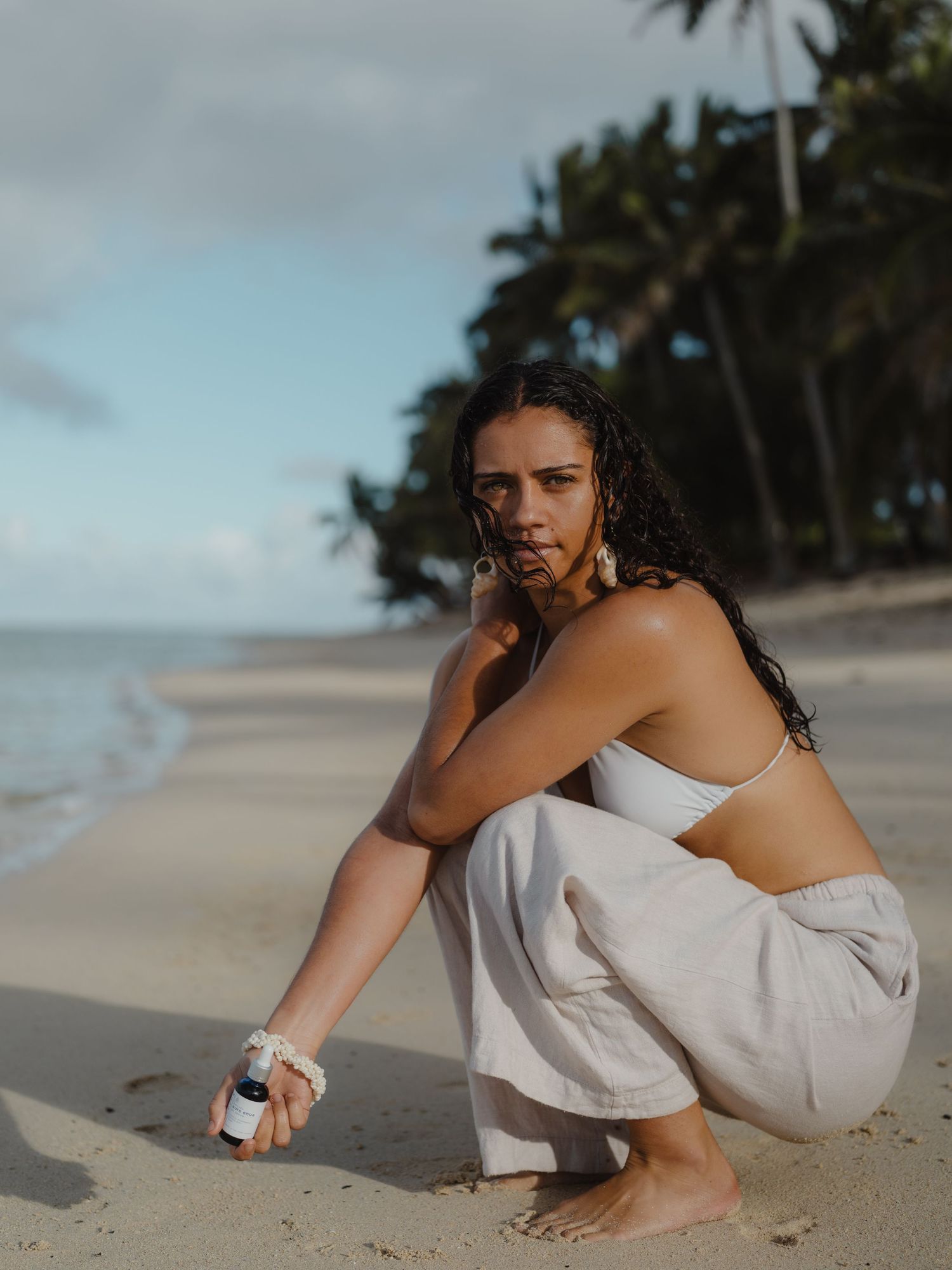 Woman crouching on a tropical beach holding a small bottle, wearing white linen pants and a bandeau top.
