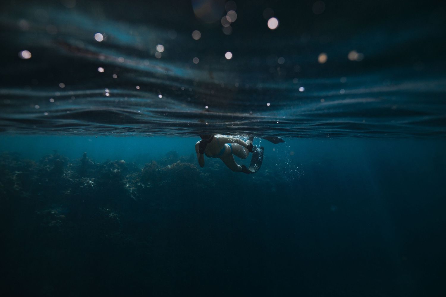 Snorkeler swimming underwater in dark blue ocean with light bubbles and bokeh effect.