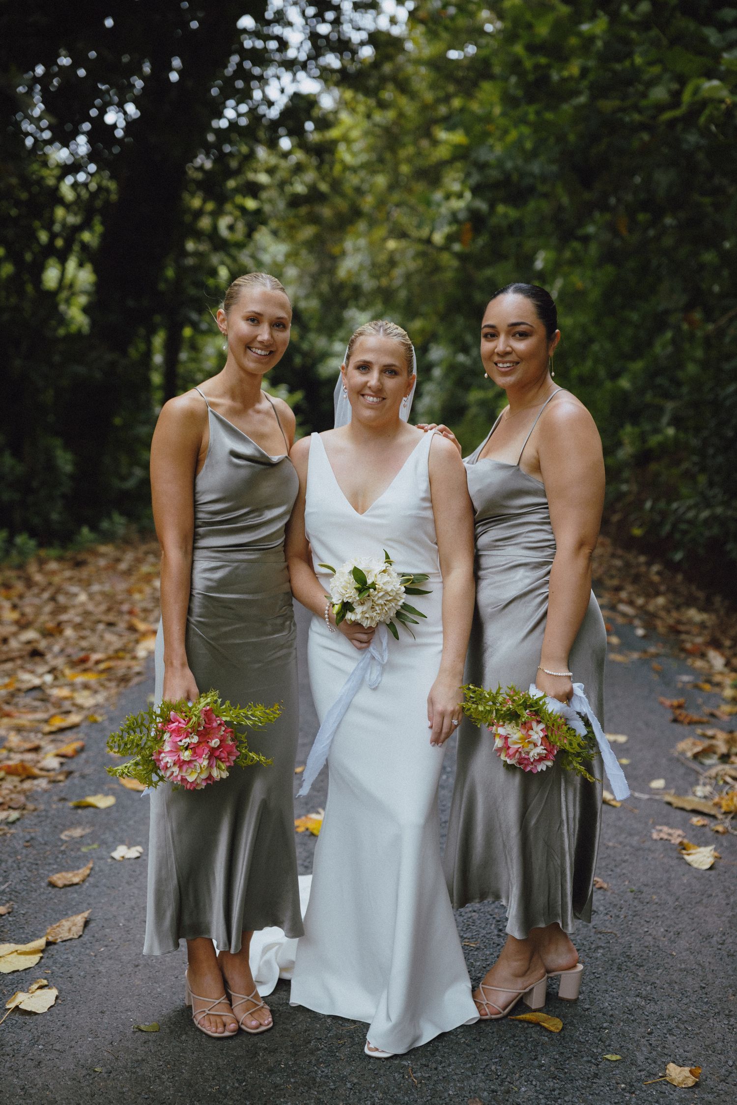 Bride in white gown with two bridesmaids in sage satin dresses holding bouquets on a tree-lined path.