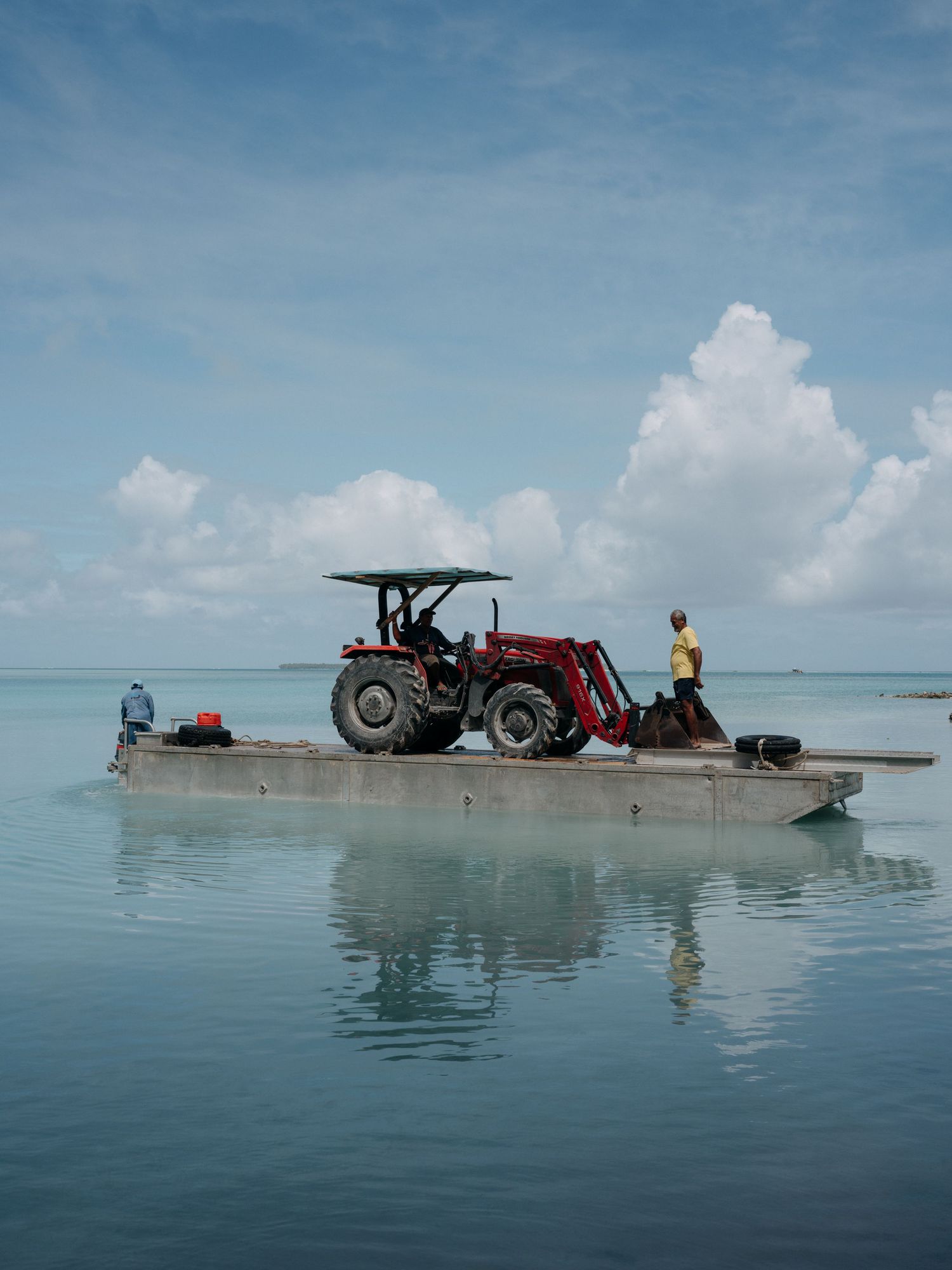 Red tractor on a flat barge floating in calm turquoise waters with workers nearby under a partly cloudy sky.