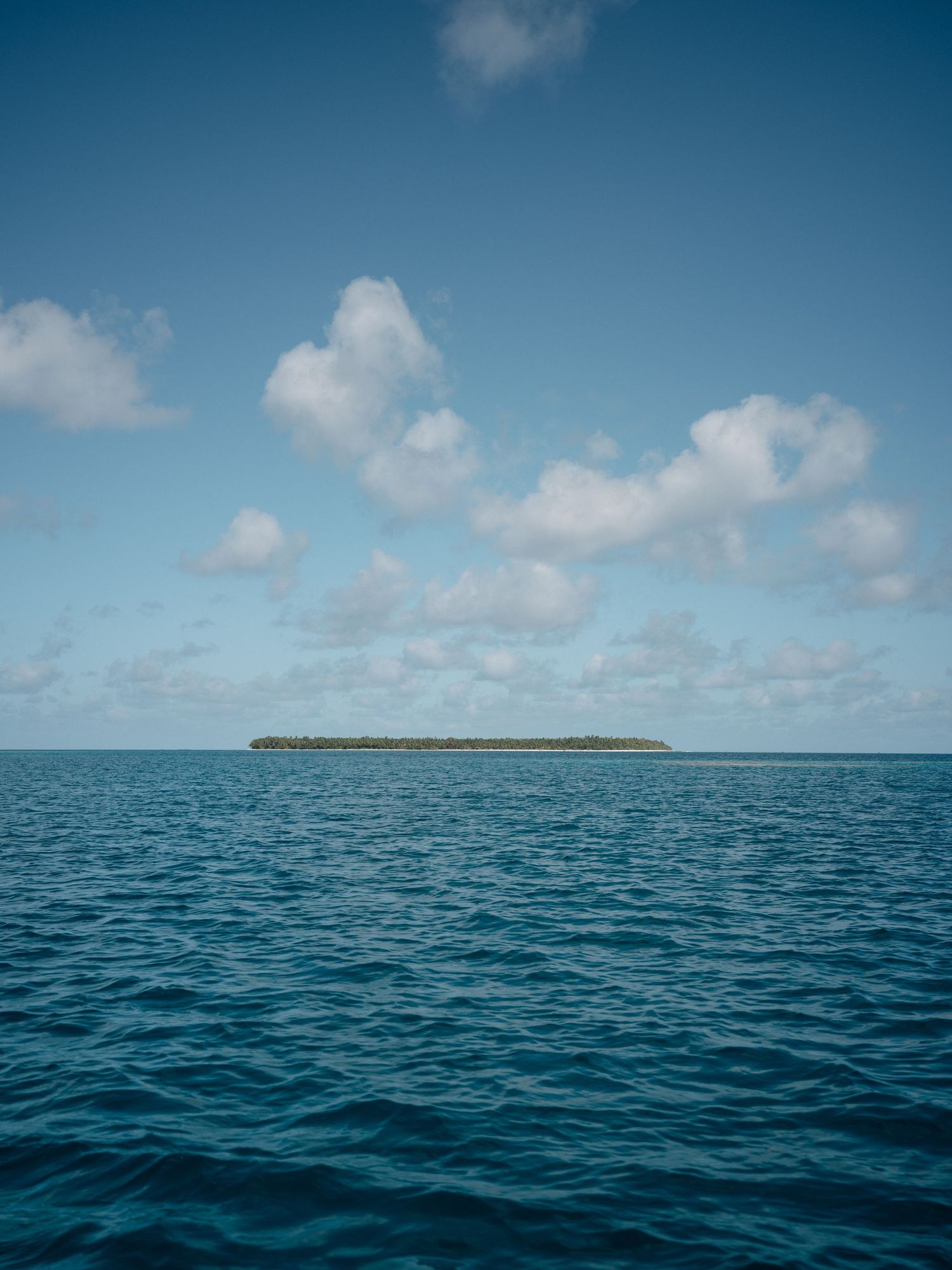 A small tropical island with lush green trees sits on the horizon of a calm blue ocean under a partly cloudy sky.