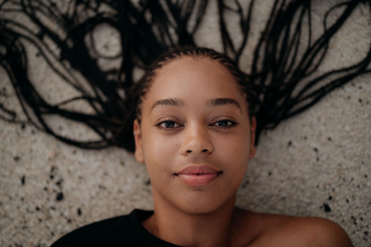 Young Black woman with long braids lying on concrete surface, looking up at camera with a calm expression.